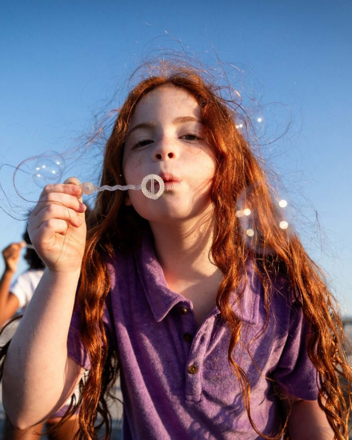 A cheerful girl with curly red hair blows bubbles in a violet polo dress, capturing playful summer moments.