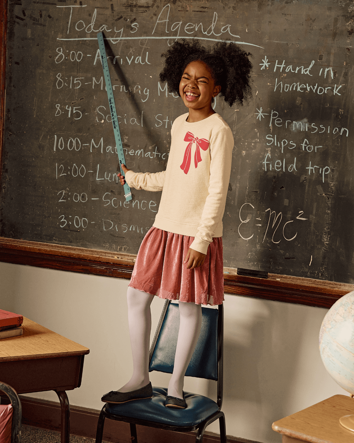 Happy girl in cream sweatshirt dress with pink bow, showing off her style in a playful classroom setting.