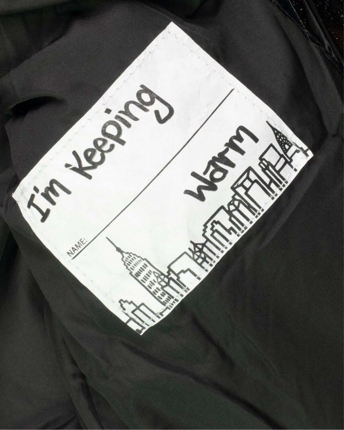 Playful label inside the Black Glitter Puffy Coat, featuring a fun ‘I’m Keeping Warm’ message with a city skyline drawing.
