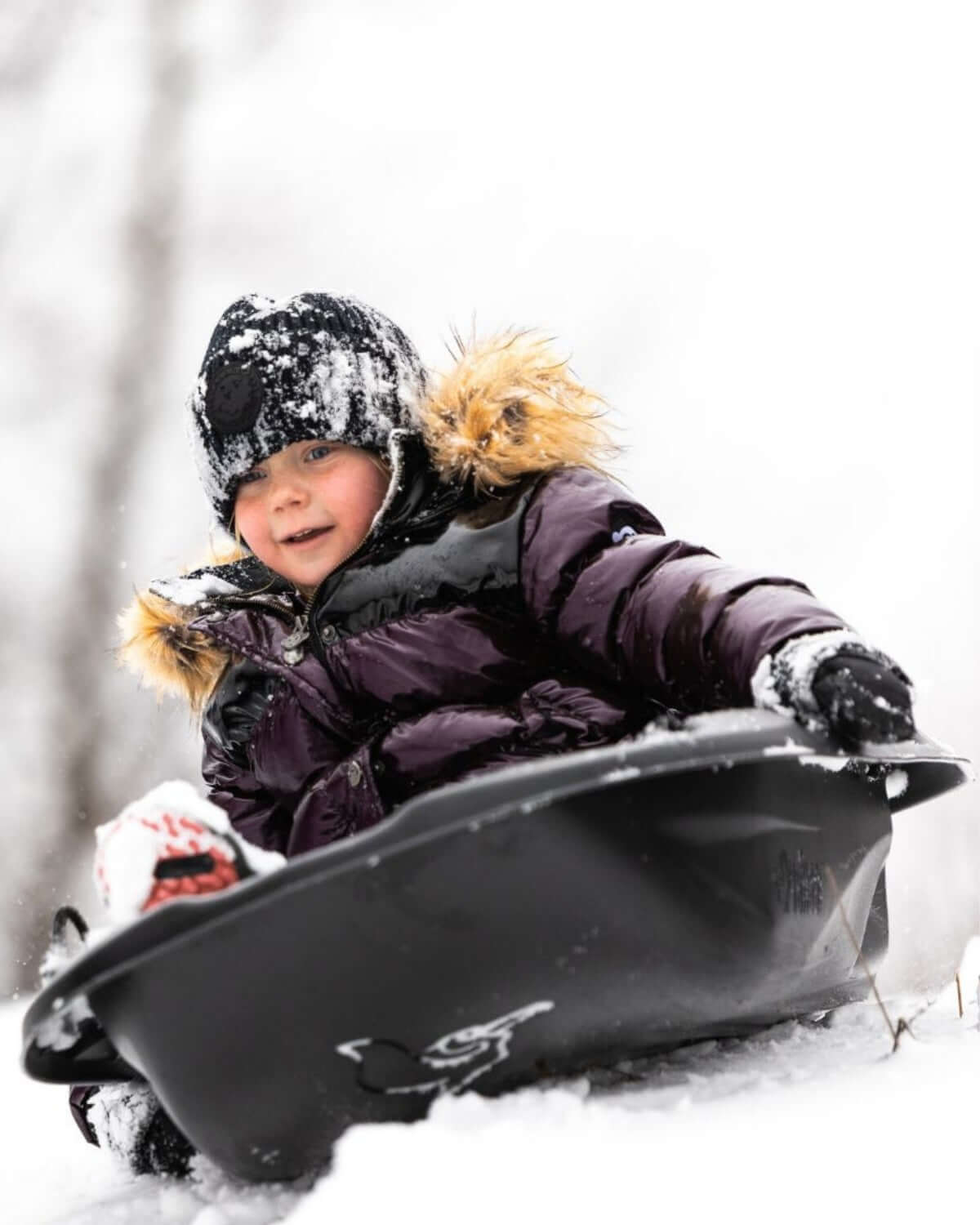 Joyful child in a burgundy puffer coat sledding through fresh snow, embracing winter fun in stylish kids clothes.