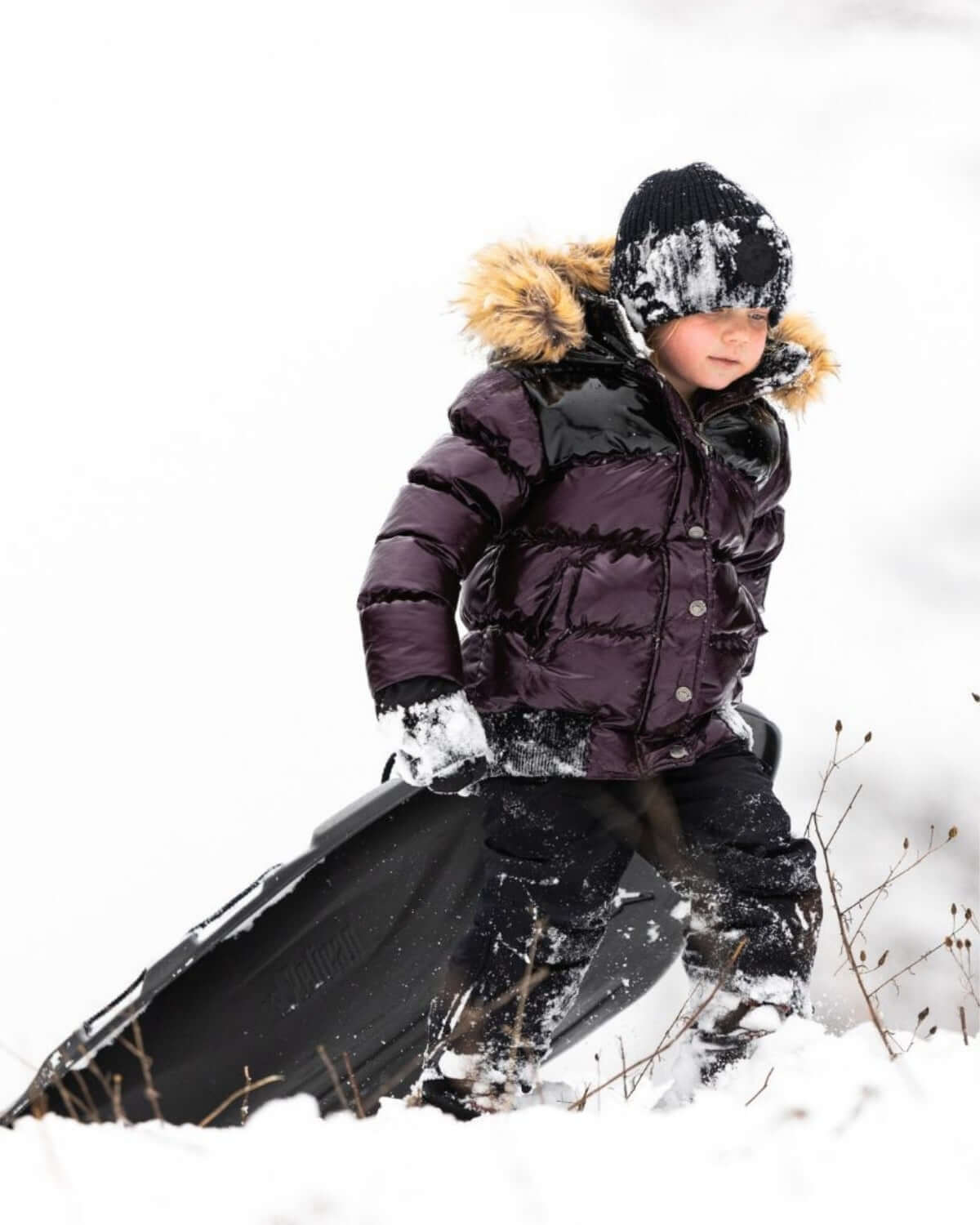 A stylish kid in a burgundy puffer coat with a faux fur hood, sledding joyfully through the snowy landscape.