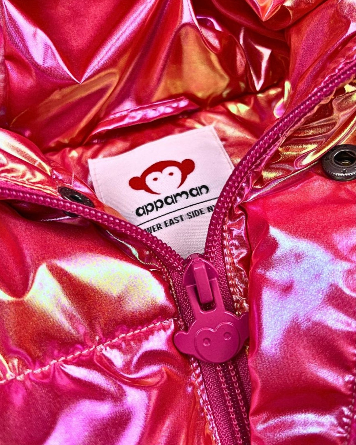 Close-up of Appaman's Citrus Sparkle Puffy Coat, featuring shiny pink fabric and playful monkey logo—fun, stylish kids' outerwear!