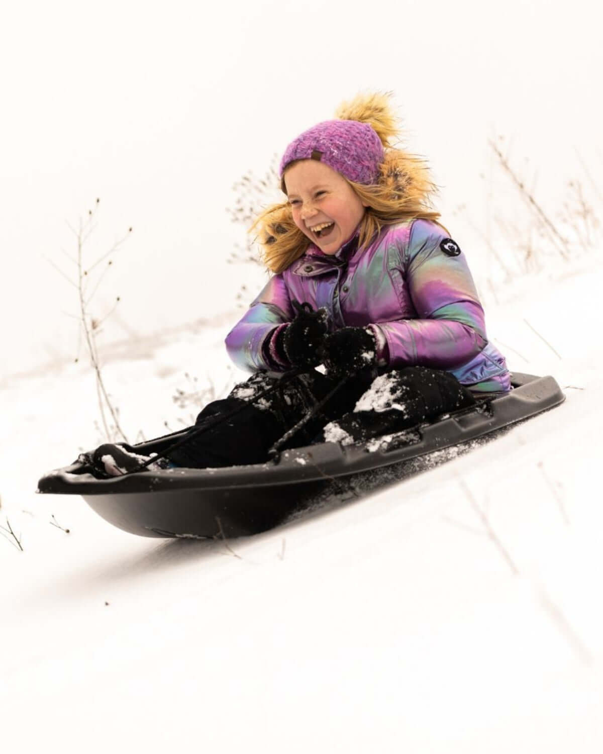 A joyful girl in a colorful puffer coat speeds down a snowy hill on a sled, showcasing high-quality kids' winter clothes.