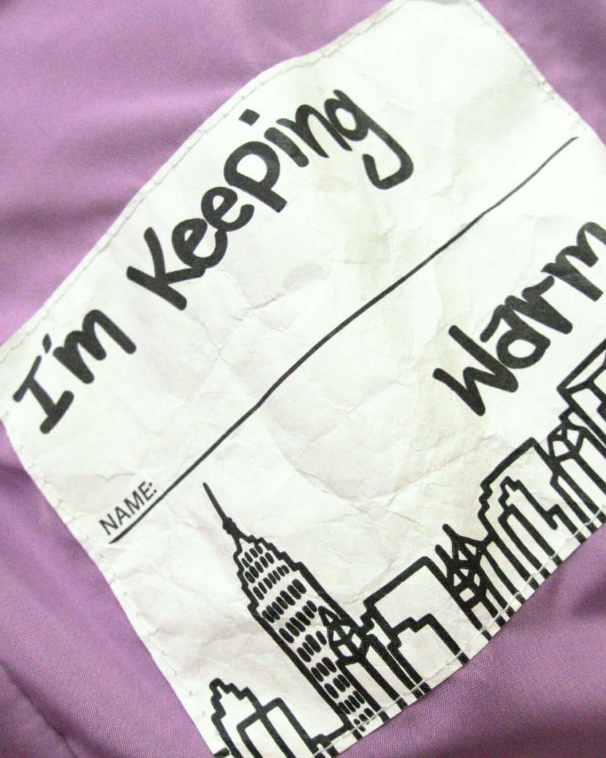 Close-up of the playful label on Glacial Purple Puffy Coat, reminding kids to stay warm in style!