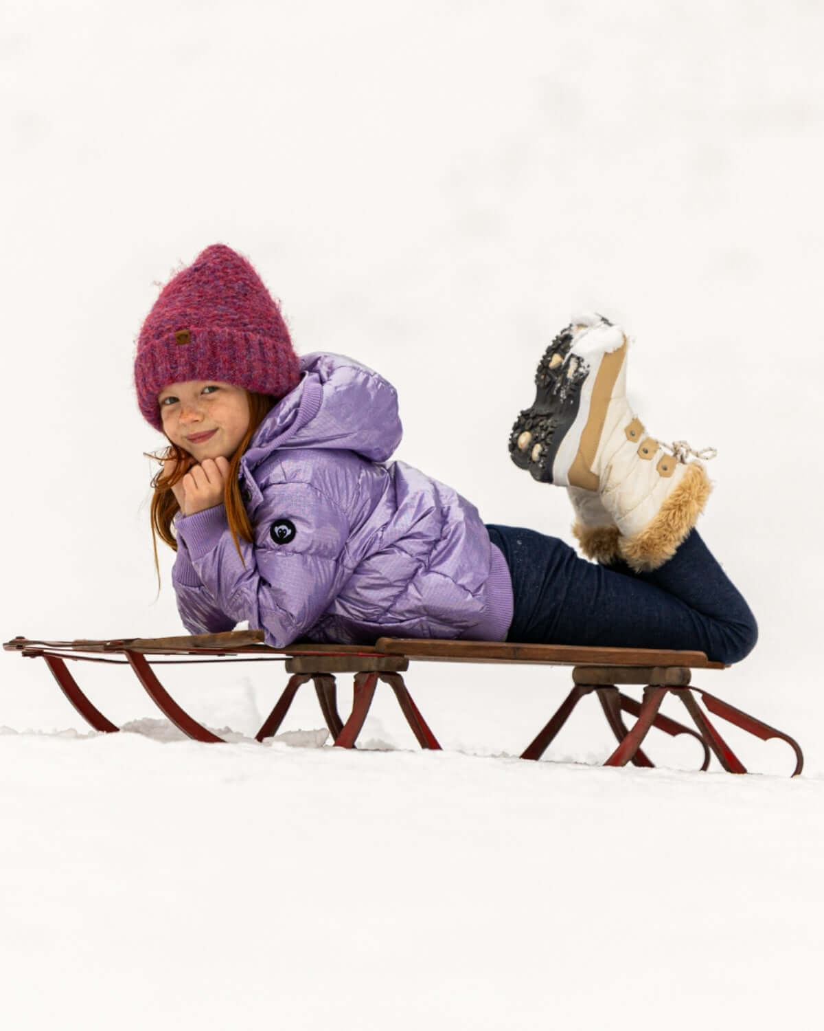 Joyful girl in a Glacial Purple Puffy Coat, cozy on a sled, embracing winter adventures in stylish kids clothes.