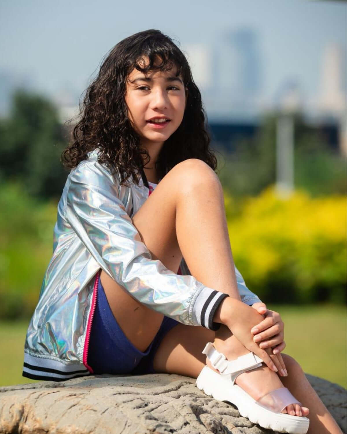 Girl wearing an Iridescent Nikki Bomber Jacket, sitting on a stone in a sunny outdoor setting.