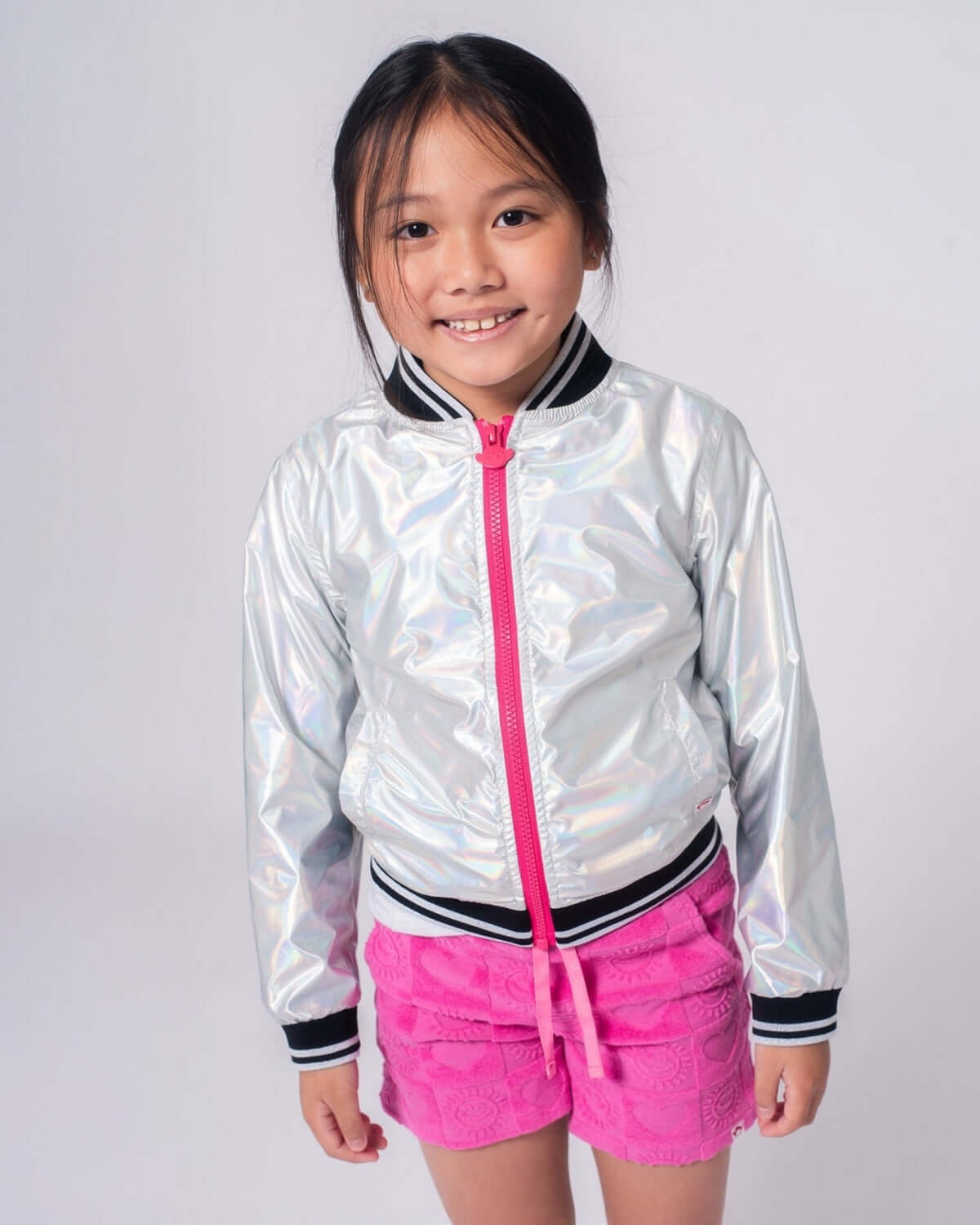 Young girl wearing an Iridescent Nikki Bomber Jacket over pink shorts, showcasing its shiny and stylish design.