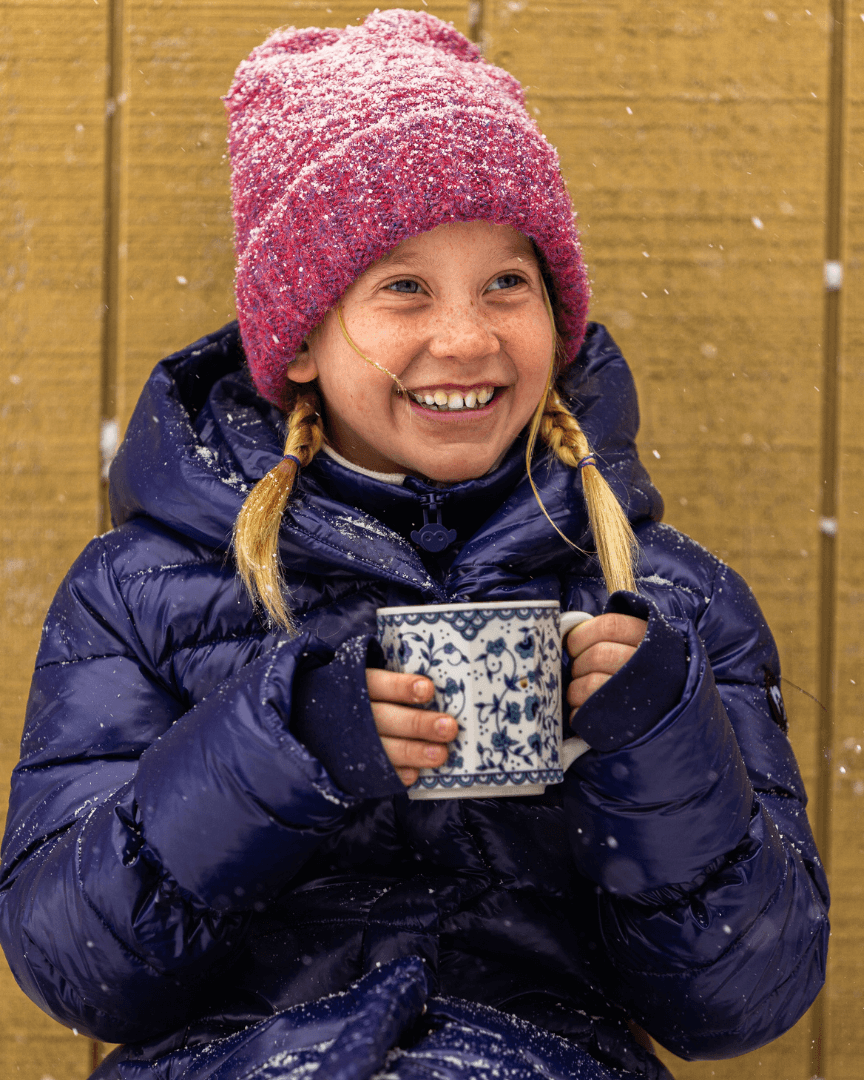 Joyful girl in a blue long puffer coat, sipping cocoa in the snow—stylish warmth for kids this winter!