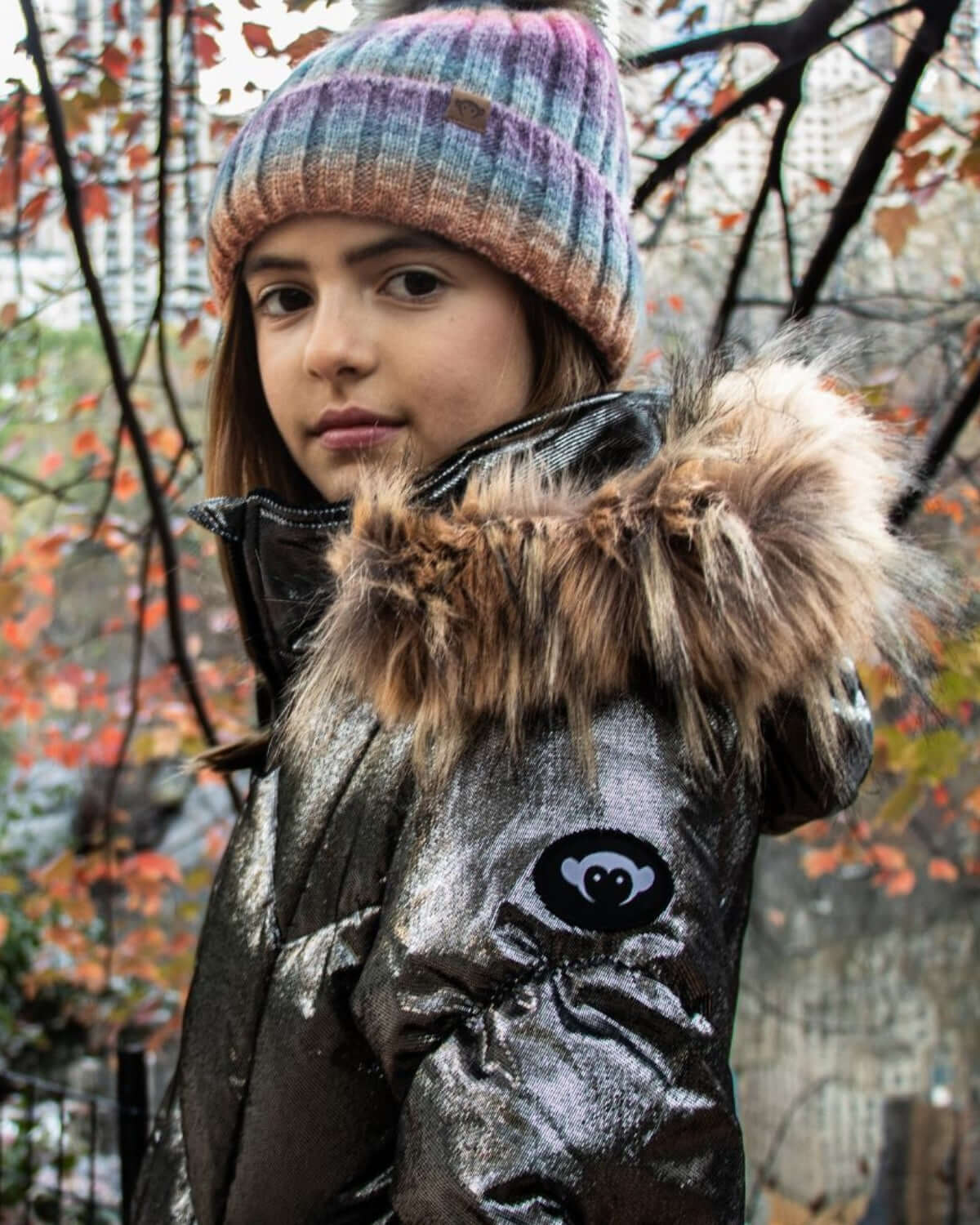 Trendy girl in shiny Moon Stone Nova Long Coat with faux fur hood, rocking a colorful knit hat in a vibrant autumn scene.