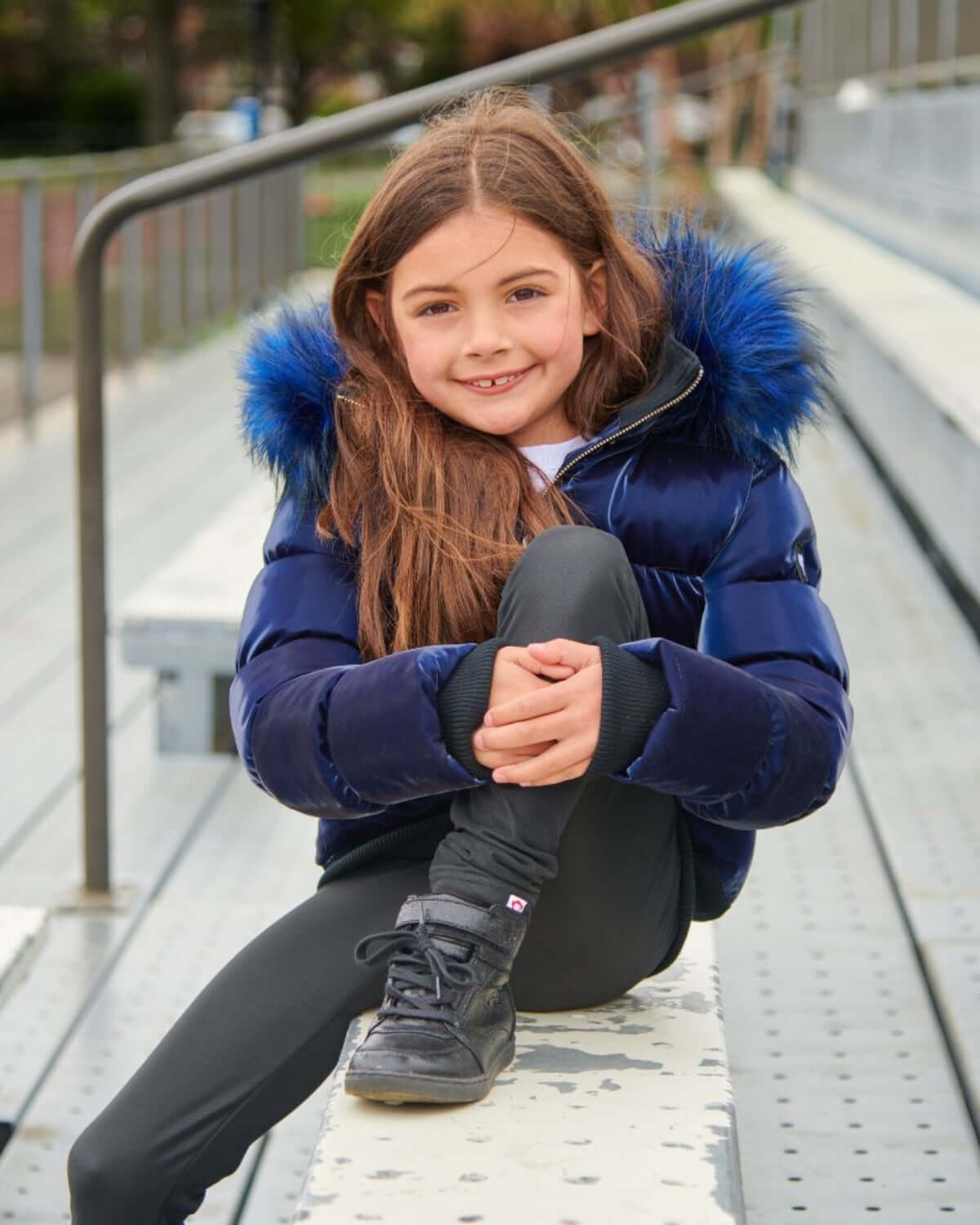A stylish girl in a navy shine Kyla puffer coat with faux fur hood, sitting on bleachers, ready for winter fun.