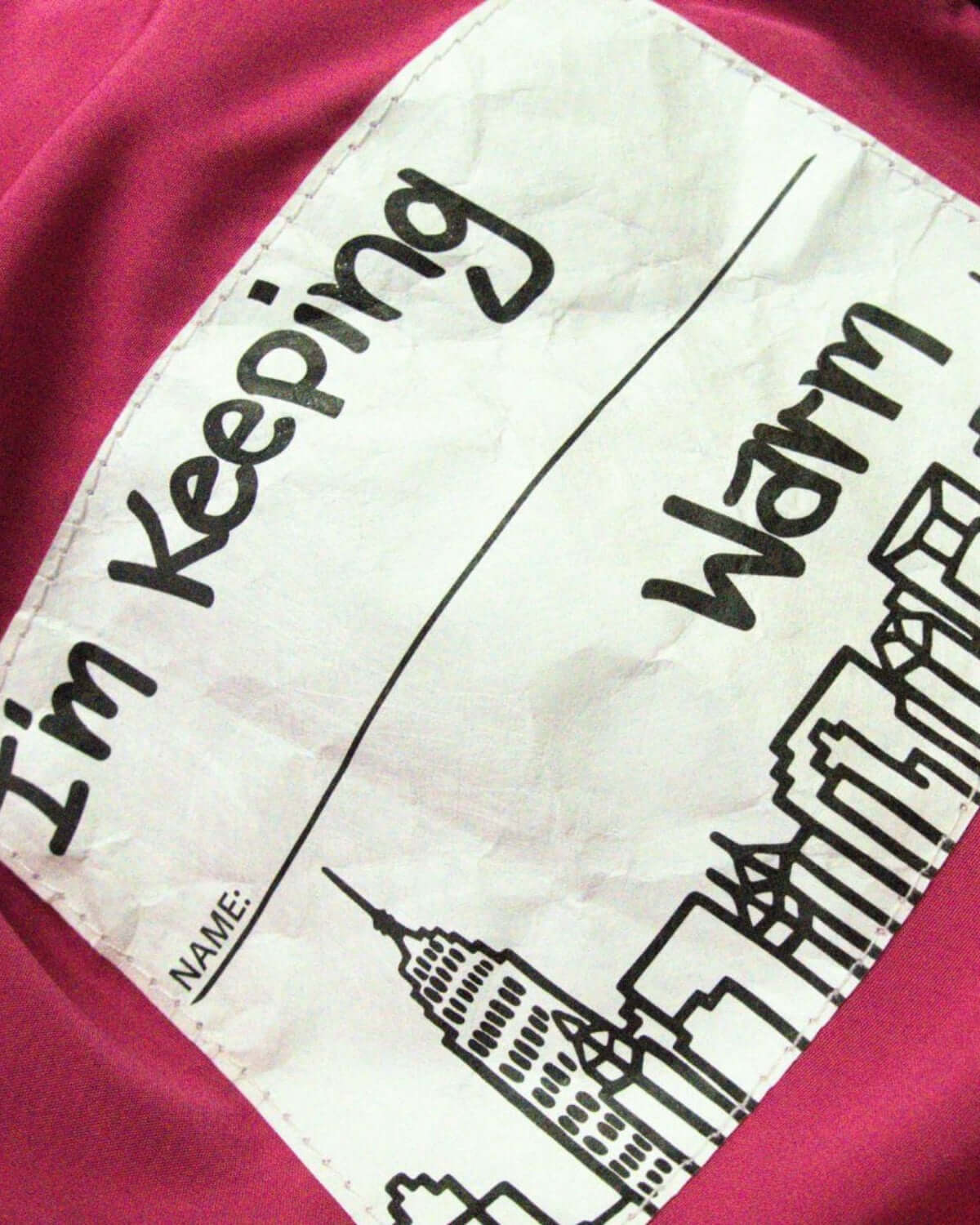 Colorful interior label of a kids' coat, playfully stating "I'm Keeping Warm" with a city skyline design.