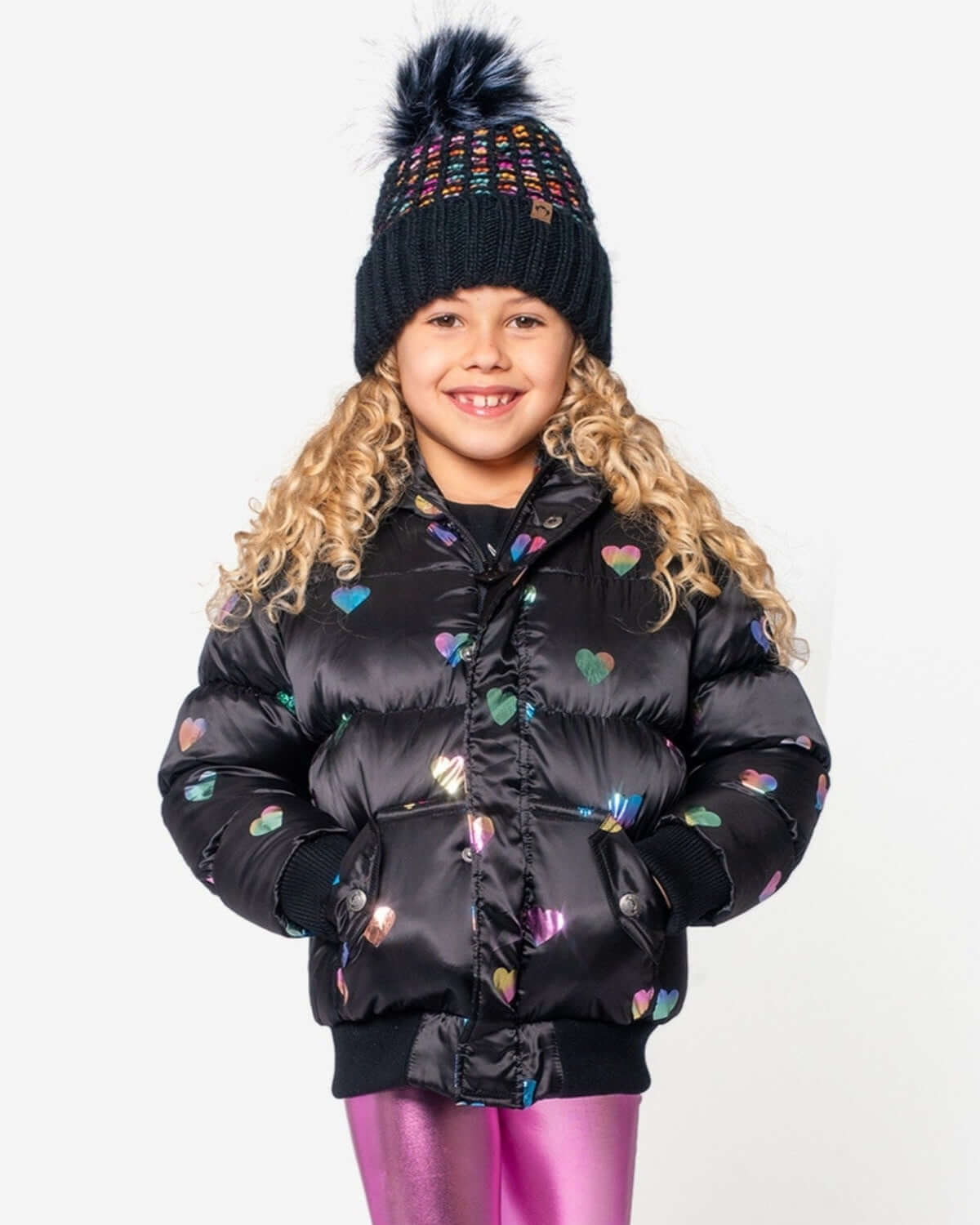 Stylish girl in a Rainbow Hearts Puffy Coat and colorful pom-pom hat, ready to conquer winter in high-quality kids clothes.