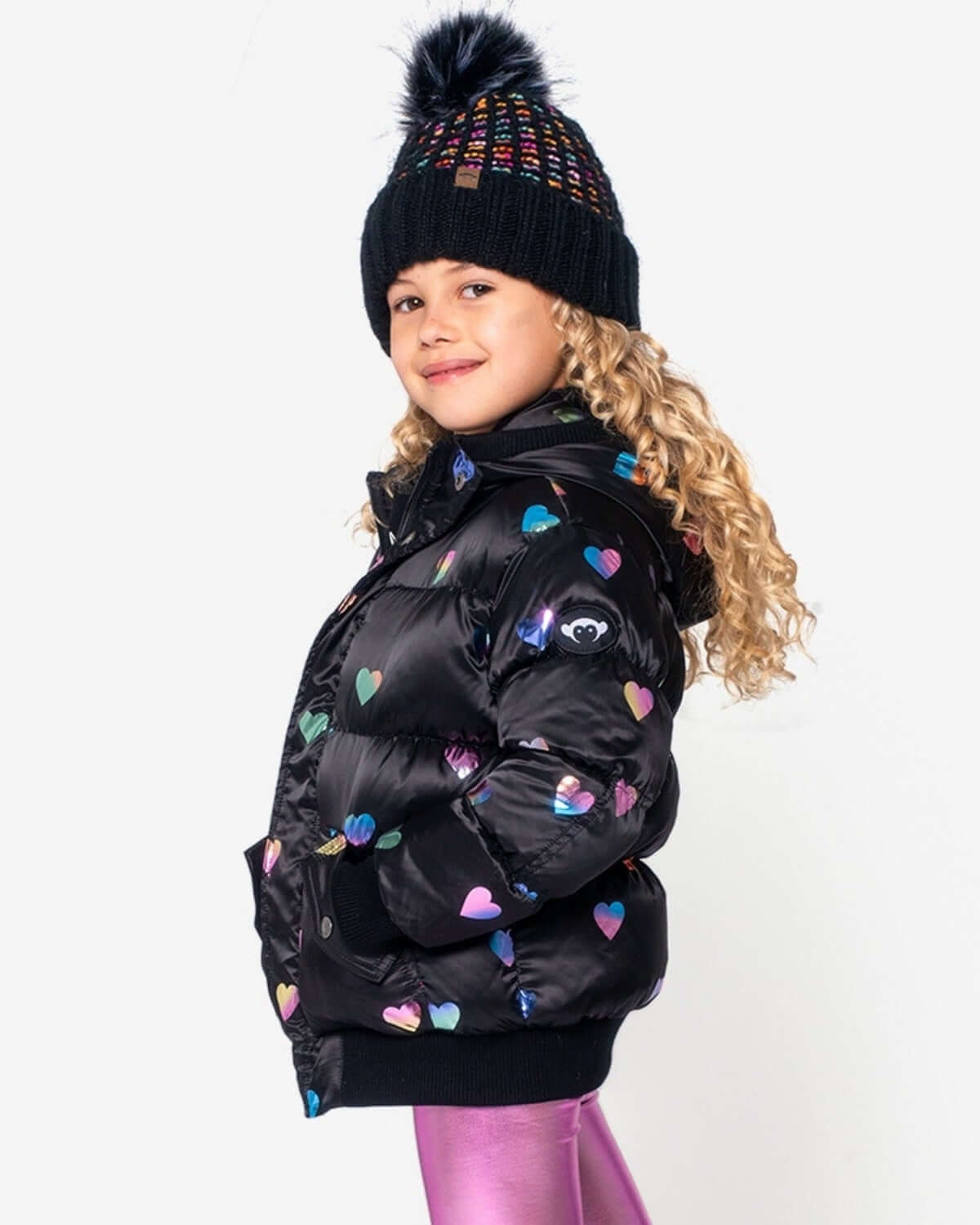 Stylish kid in rainbow hearts puffy coat and playful beanie—perfect for vibrant winter adventures. High-quality kids clothes!