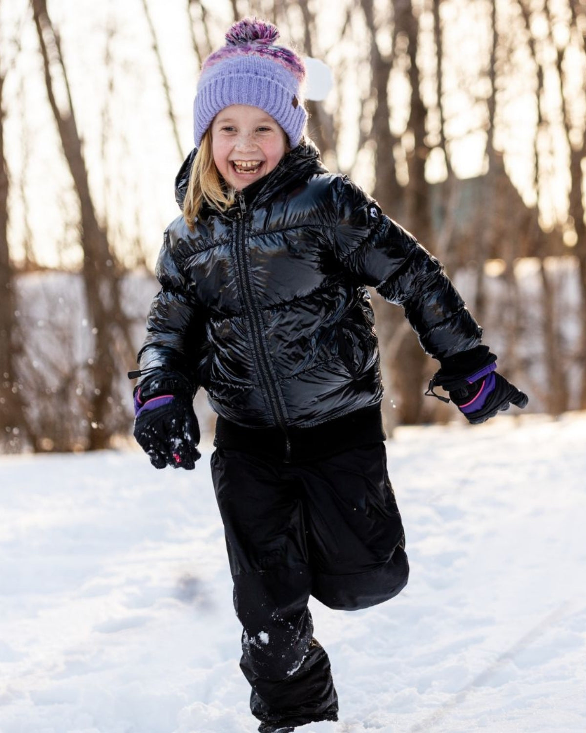 Joyful girl in a shiny black Flurry Coat and cozy hat, enjoying winter fun in the snow with style! Perfect kids' winter fashion.