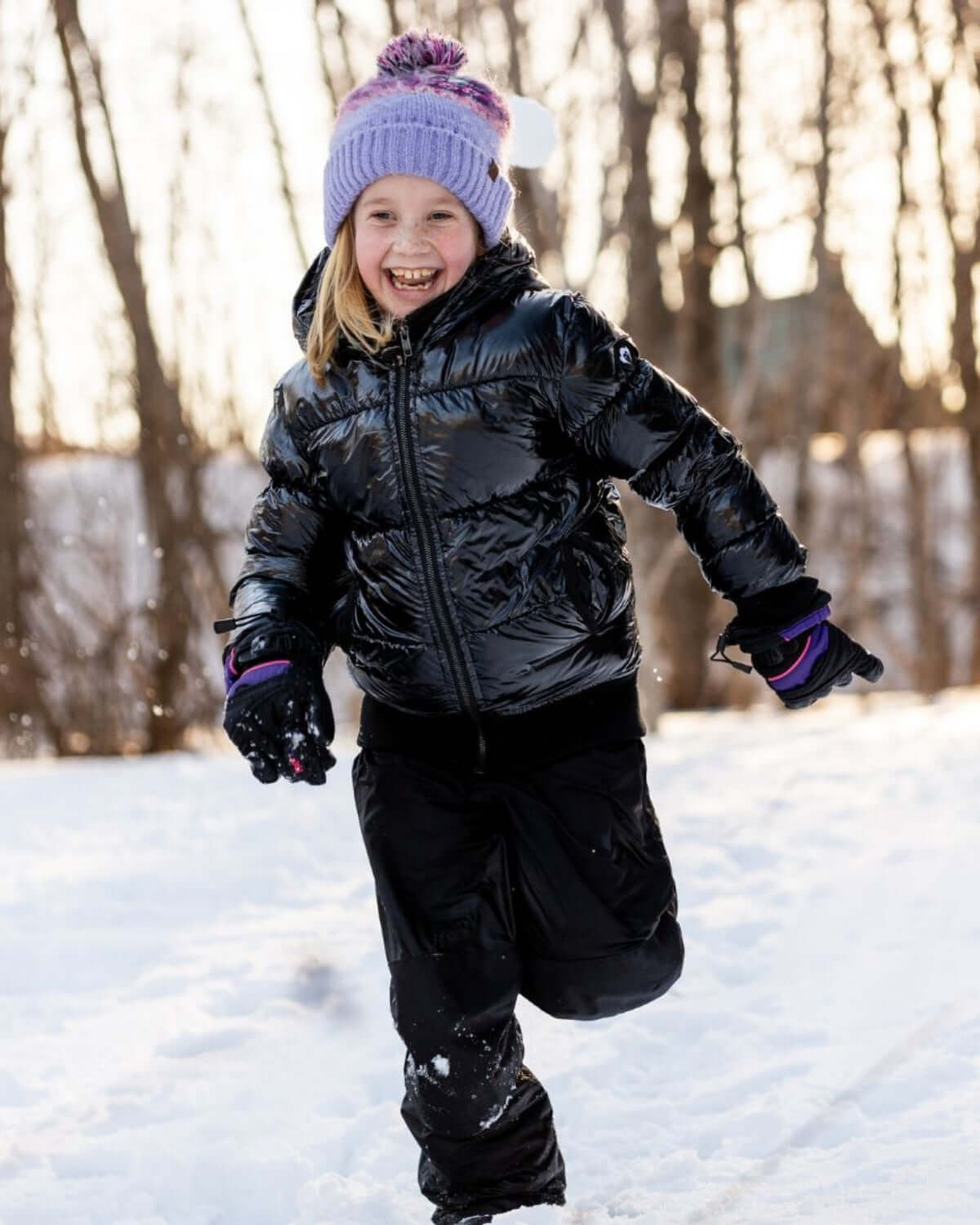 Child running in snow wearing Shiny Black Flurry Coat and winter accessories, smiling in winter landscape.