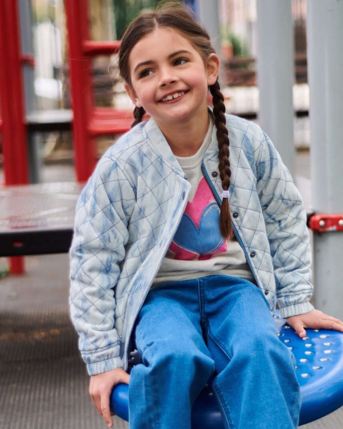 A happy girl in a trendy quilted jacket and Ruby Sweatshirt, playing on a colorful playground, showcasing kids' stylish clothes.