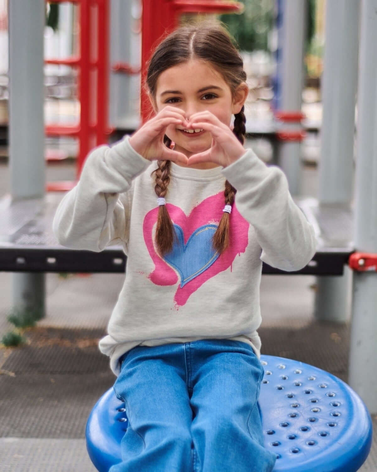 Playful girl in a Ruby Heart Sweatshirt making a heart shape with her hands, showcasing stylish kids clothes for back to school.