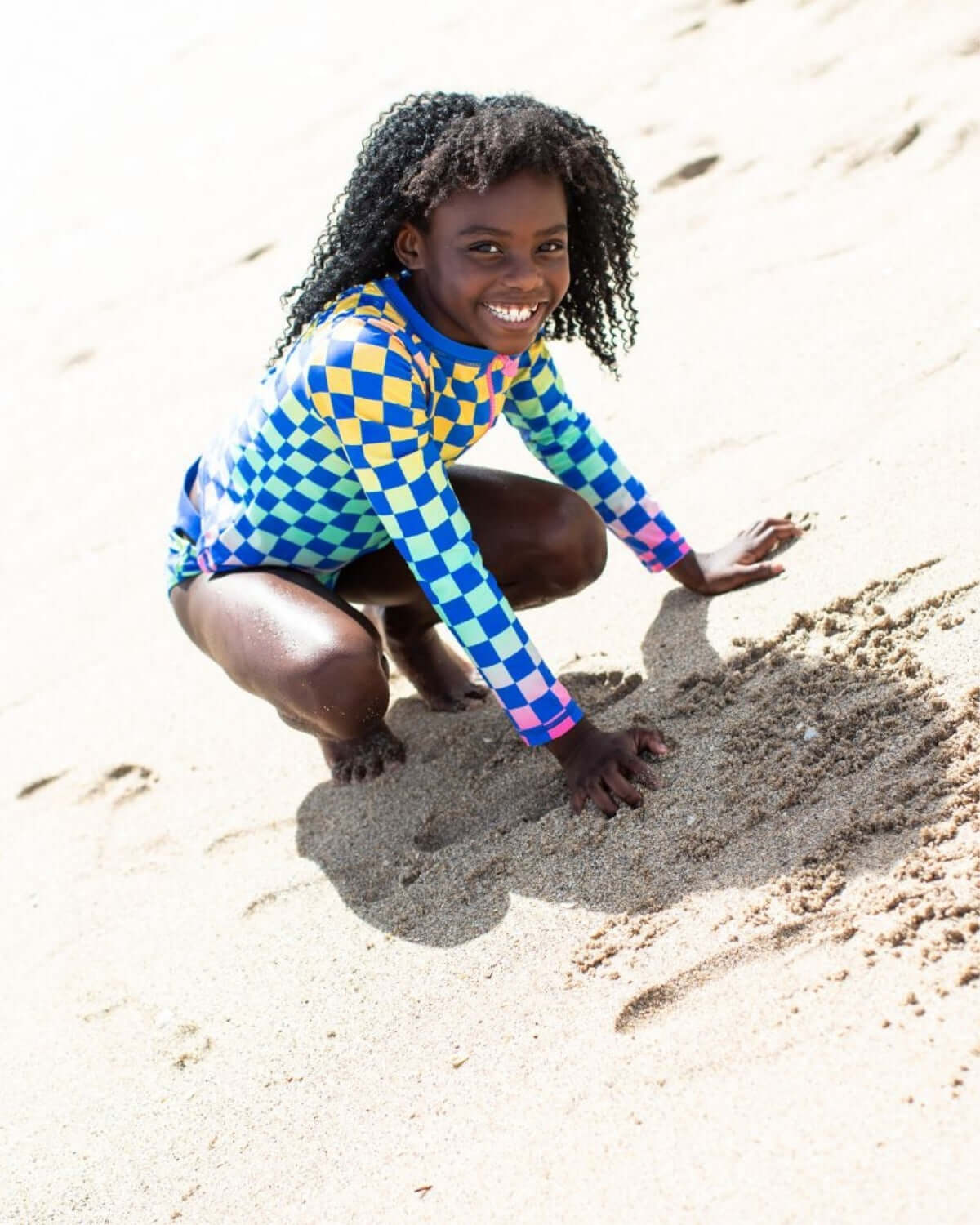 Smiling girl in a vibrant checkerboard rash guard, playing in the sand at the beach, showcasing fun and adventure.