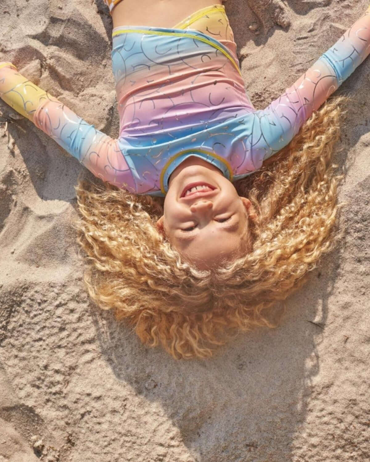 A joyful child wearing the Summer Joy Oceana Bikini, relaxing on the beach with a vibrant wrap-top and curly hair.