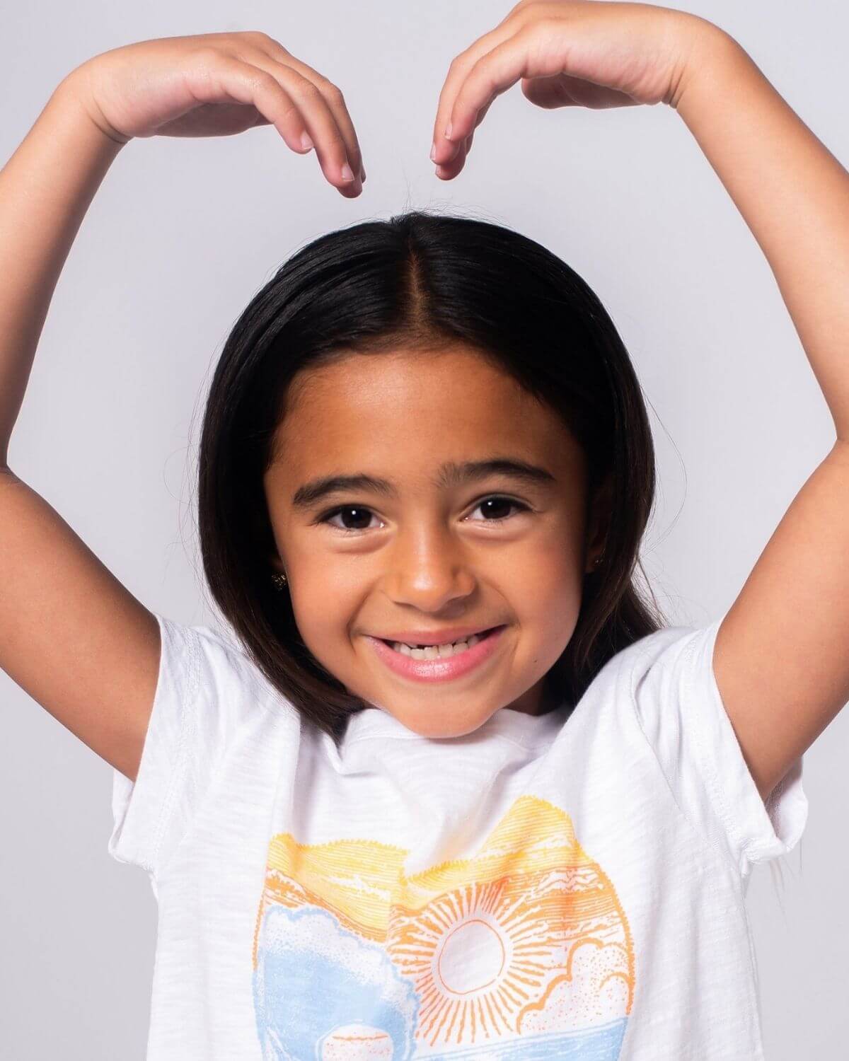 A joyful girl in a white Beach Love Circle Tee making a heart shape with her hands, ready for sunny adventures!