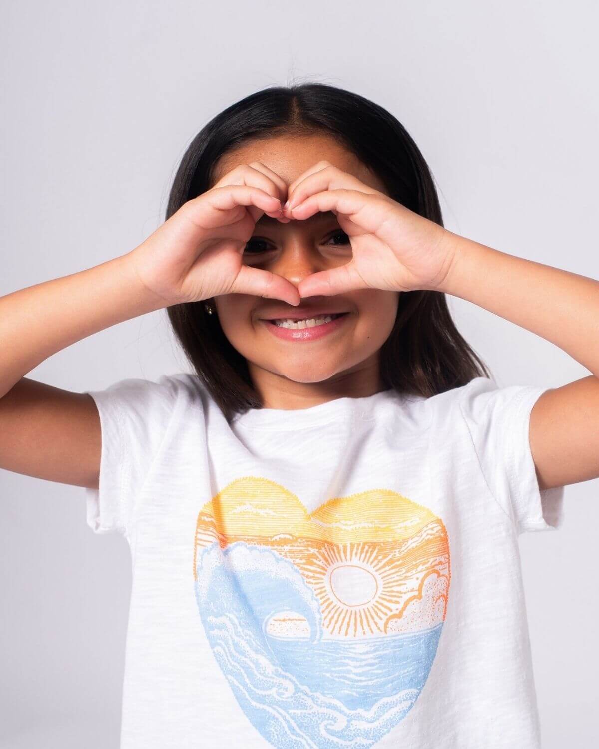 Little girl in a white Beach Love Circle Tee making a heart shape with her hands, radiating sunny vibes and joy.