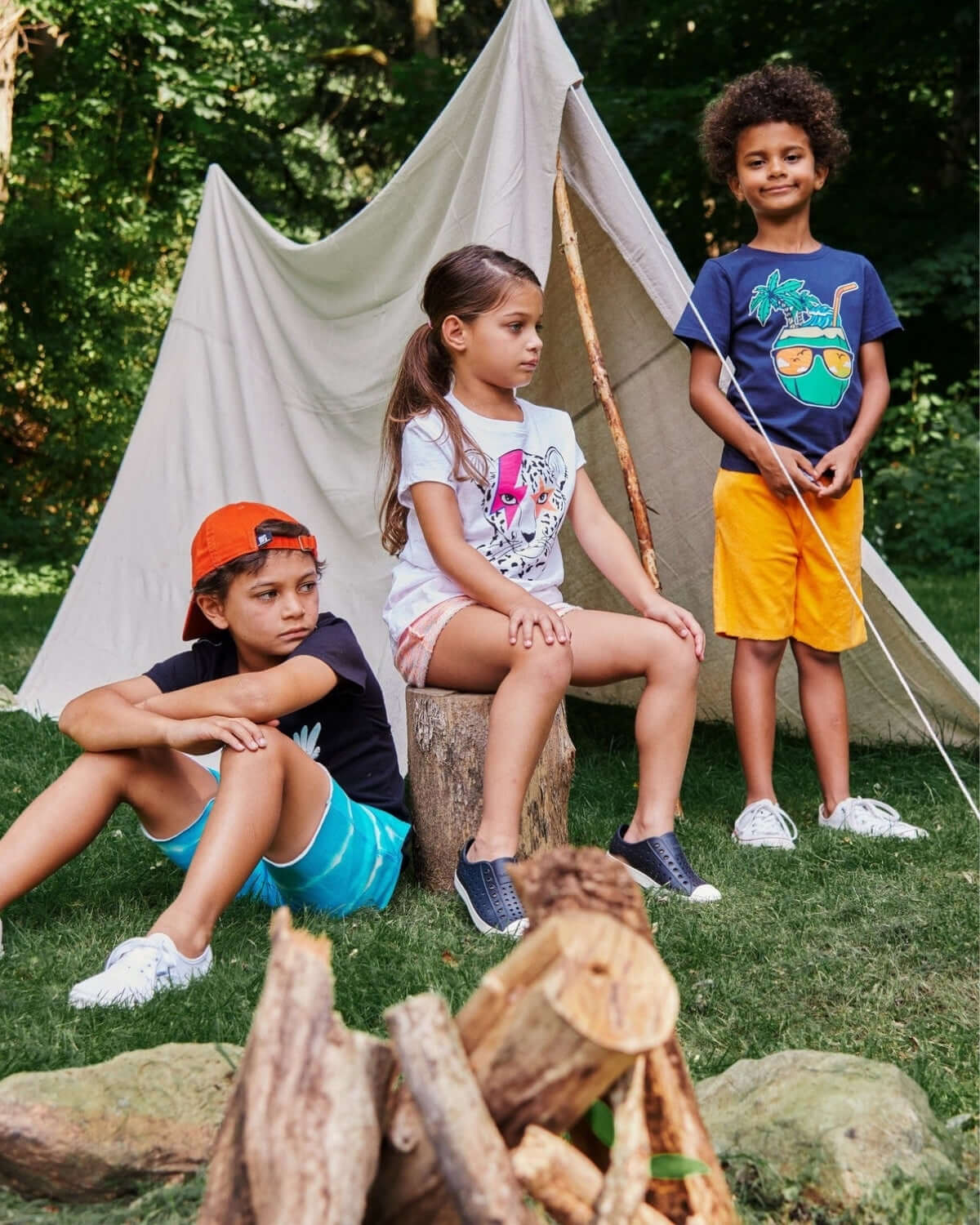 Three stylish kids in fun tees lounging by a cozy campfire, showcasing playful kids clothes perfect for back to school adventures.