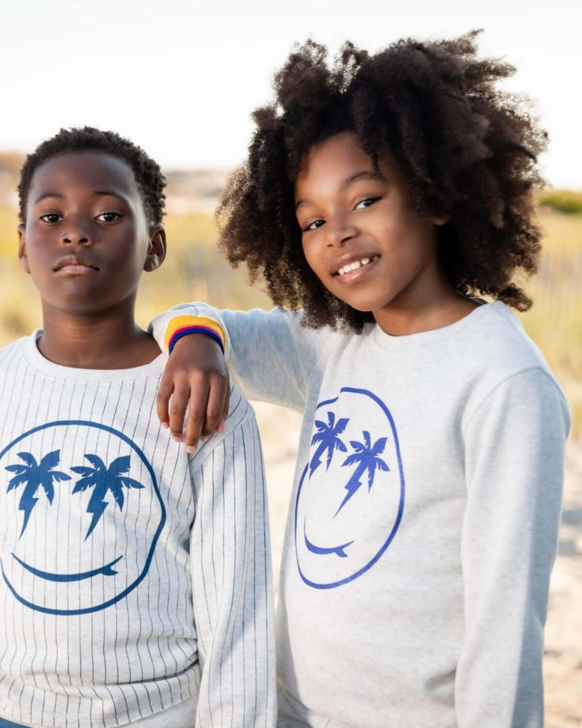 Two stylish kids in playful sweatshirts with palm tree smiles, radiating beach vibes and cozy charm.