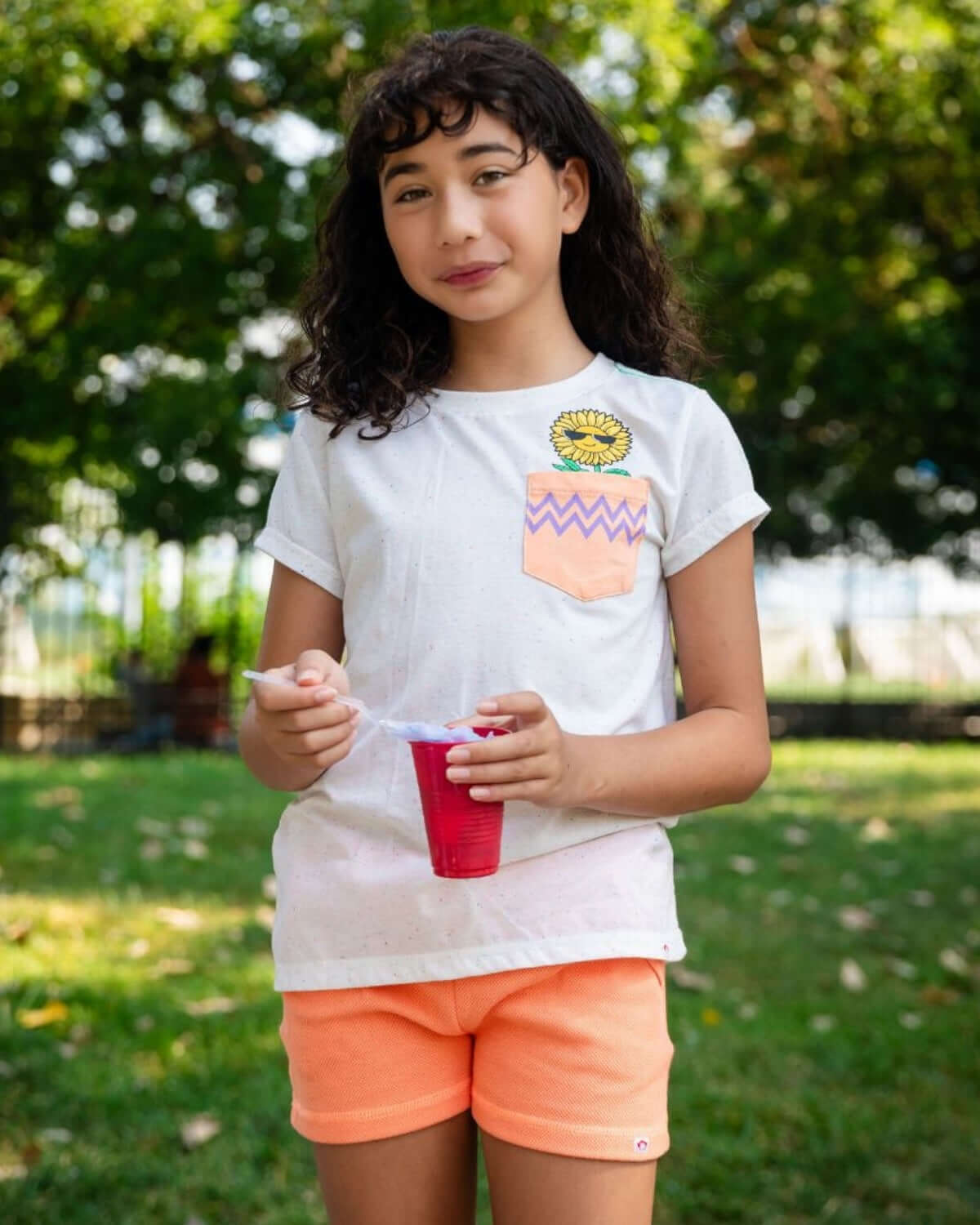 Girl wearing a Speckled White Pocket Tee with a sunflower design and orange shorts, enjoying a treat outdoors.