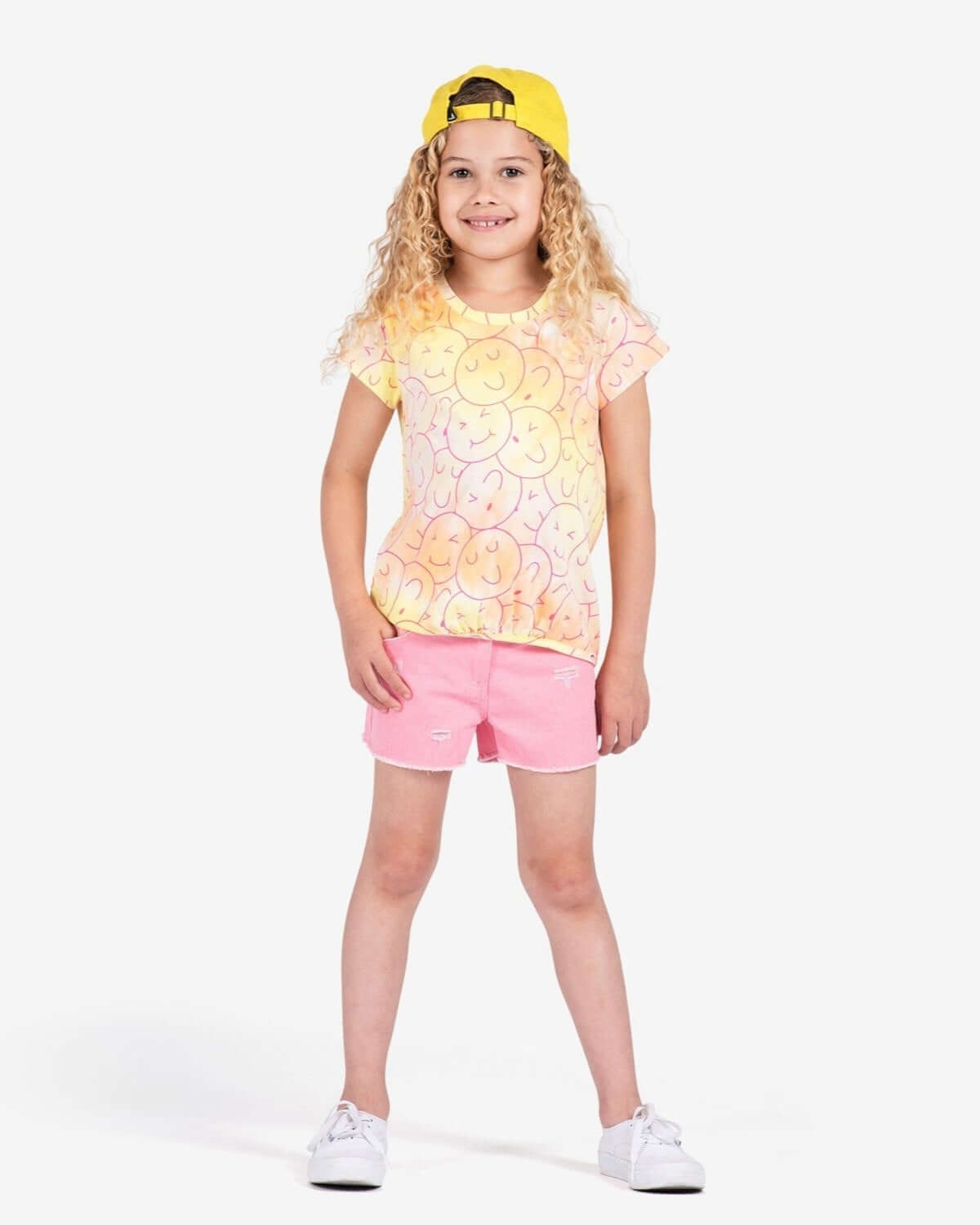 A cheerful girl in a multicolored smiley face tee and pink shorts, radiating playful vibes and fun in stylish kids clothes.