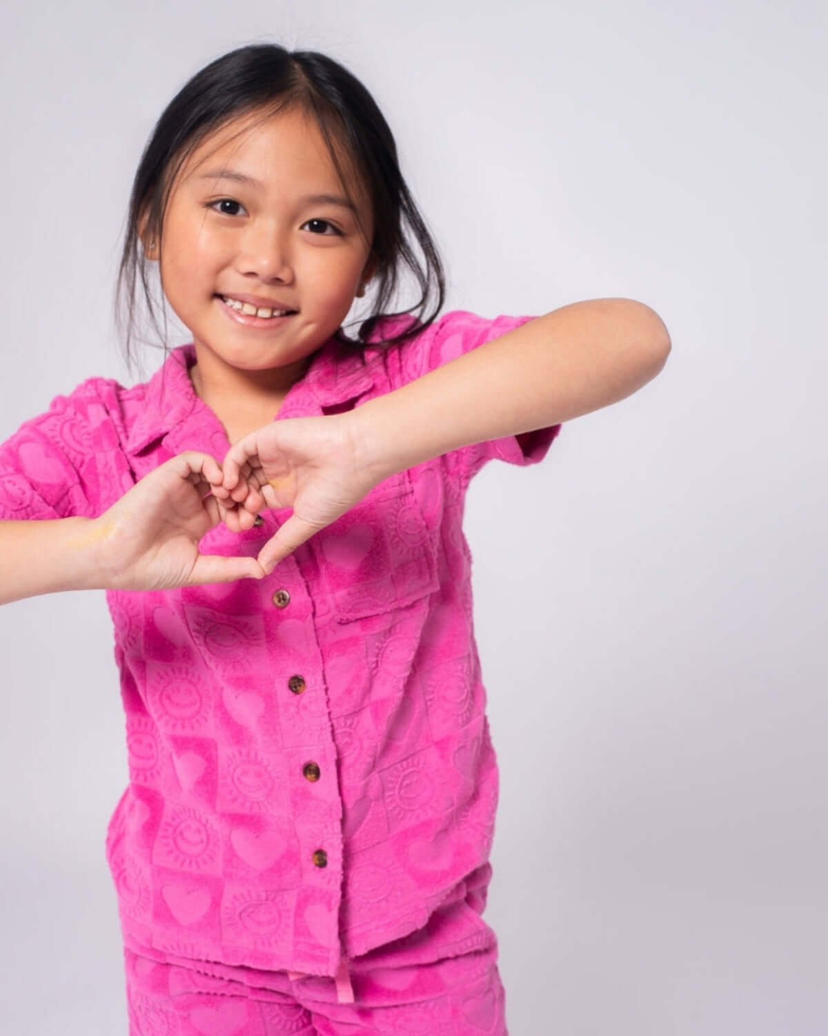A happy girl in a pink heart-patterned resort shirt shows love with a heart hand gesture, shining in stylish kids clothes.