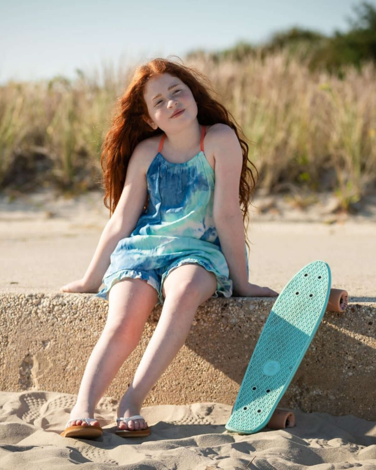 Girl sitting on the beach wearing a Turquoise Water Sanibel Tank with a turquoise skateboard next to her.