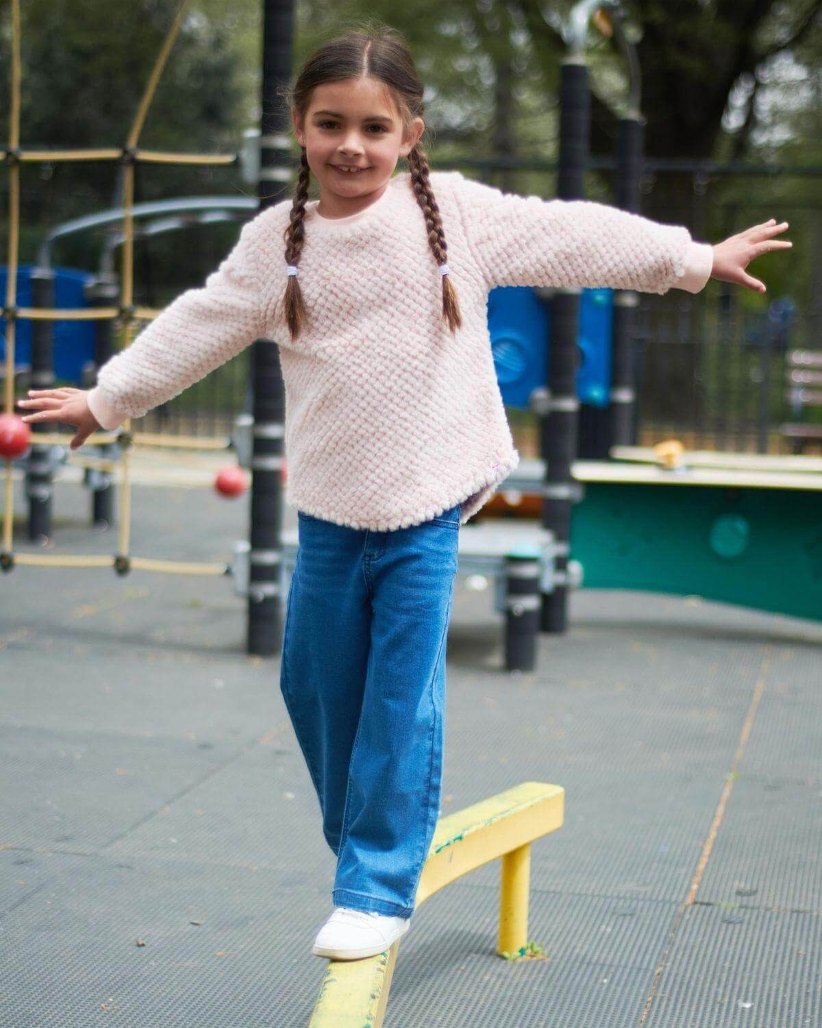 Stylish girl balancing on a playground beam in a cozy vintage pink Laurel Top, showcasing kids’ playful fashion.