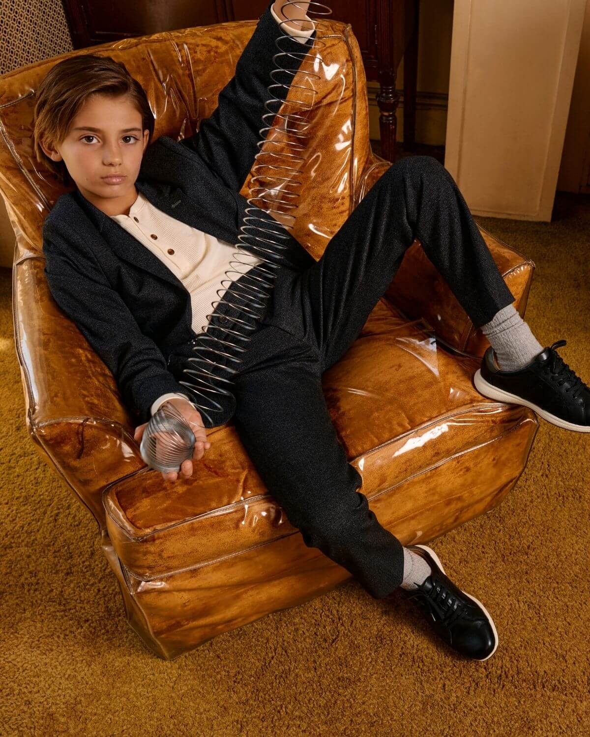 Cool kid in a black herringbone stretchy mod suit, lounging in style with a playful spring toy—effortless charm and comfort!