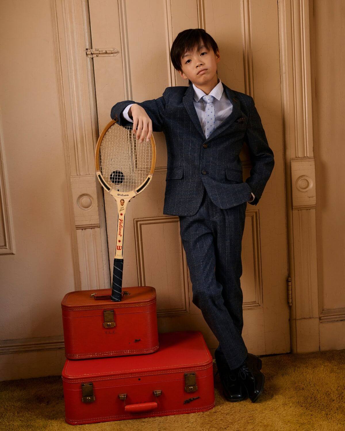 Stylish kid in Gotham Plaid Stretchy Mod Suit, posing confidently with tennis racket and vintage suitcases.