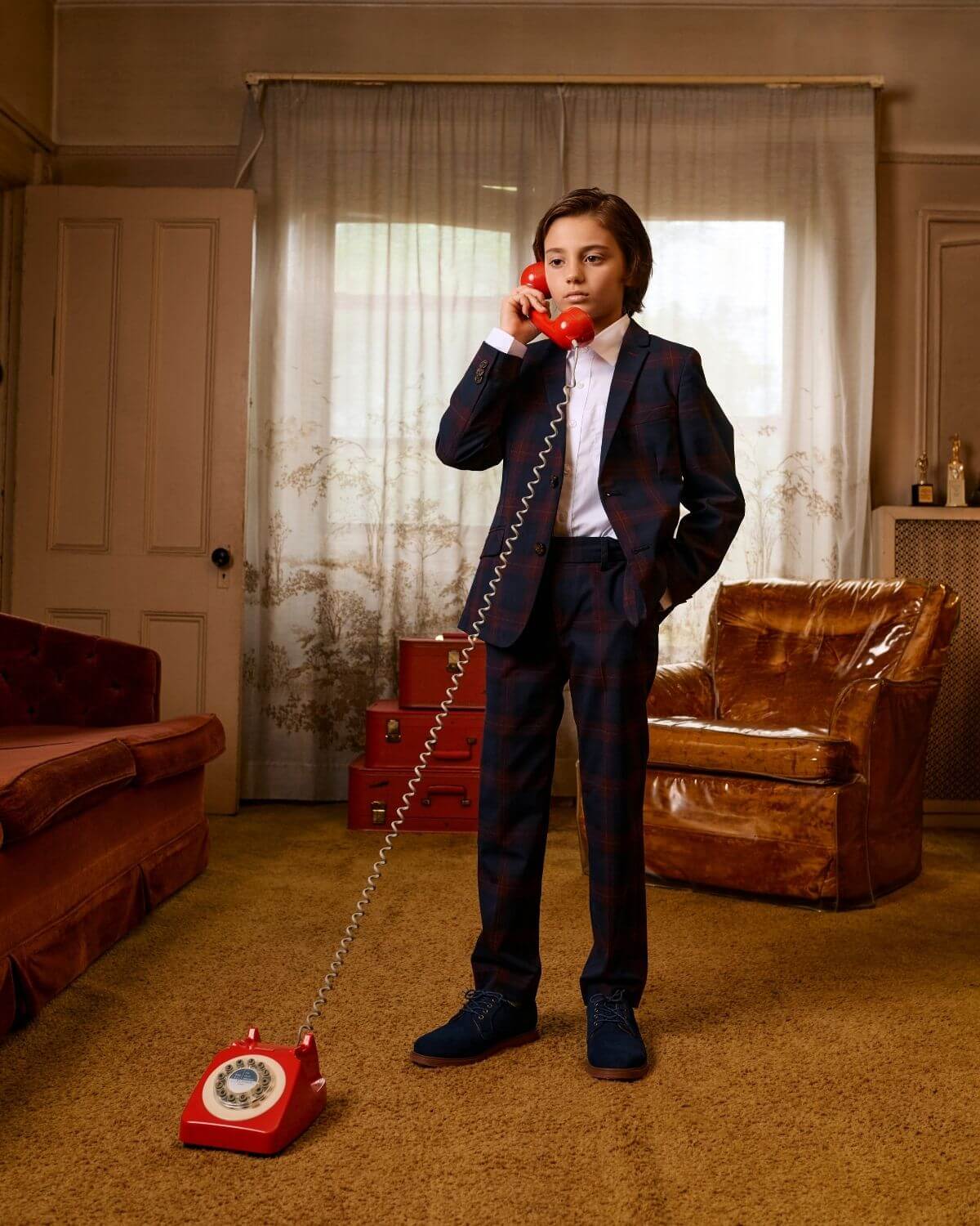 Stylish boy in Navy/Cranberry plaid suit, confidently chatting on a retro red phone in a chic vintage room.