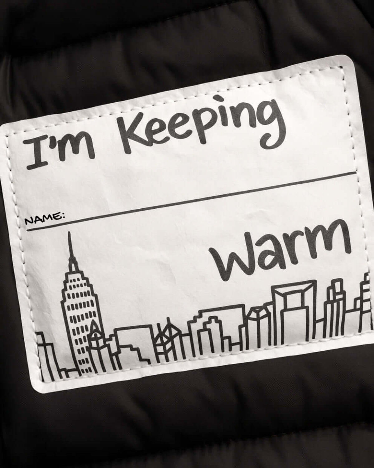 A close-up of a custom puffer jacket interior label patch that reads 'I'm Keeping Warm' with a blank 'NAME:' line and a black line-art city skyline, set against a black quilted fabric background.