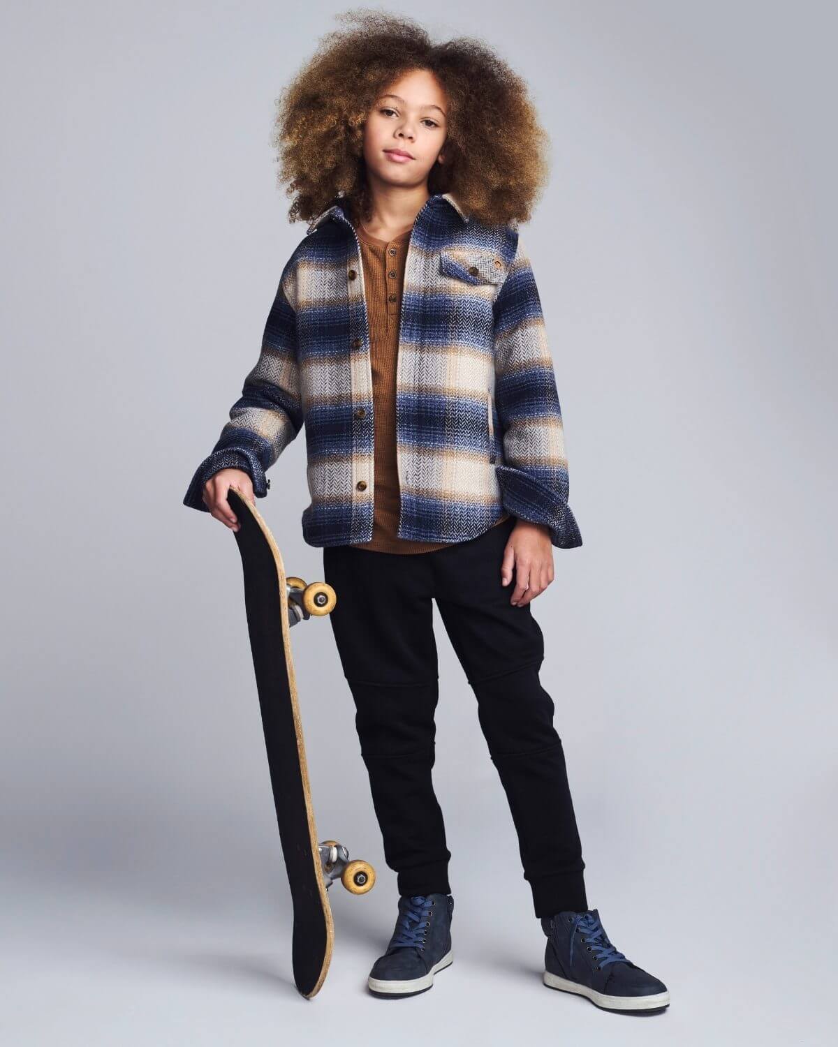 Child wearing a plaid jacket holding a skateboard on a plain background