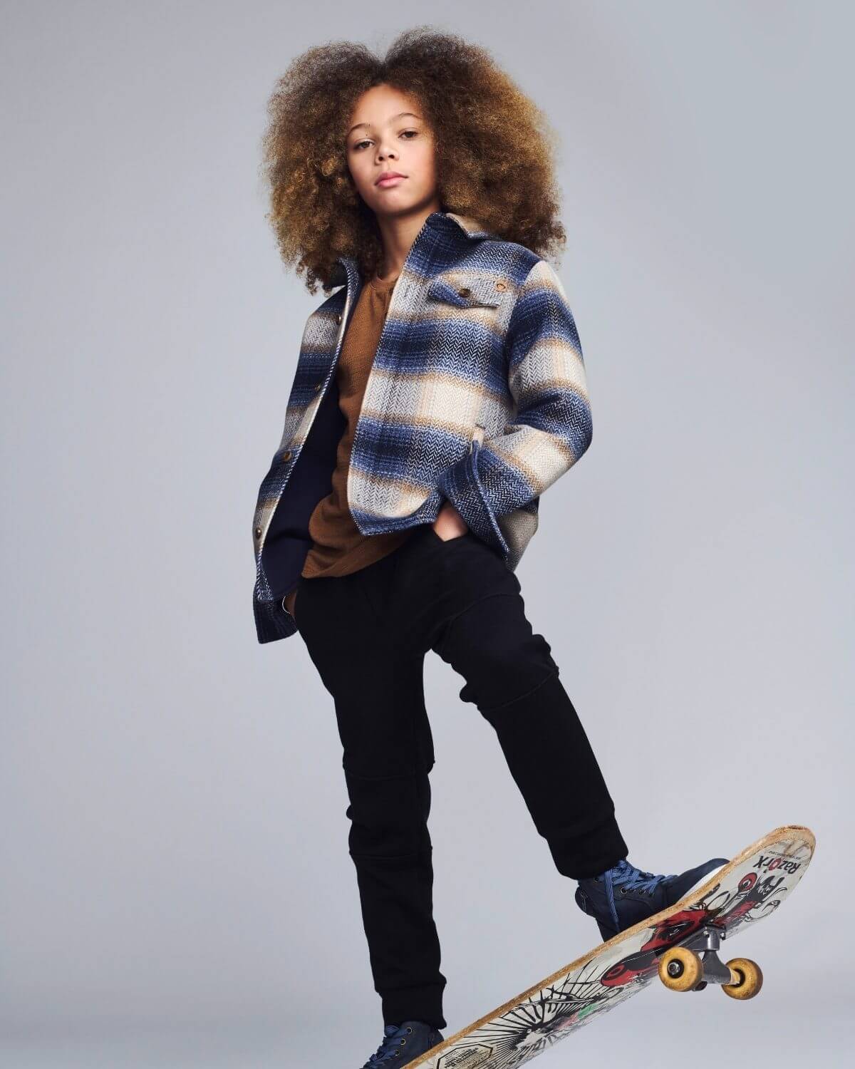 Person wearing a plaid jacket and black pants, standing on a skateboard against a gray background