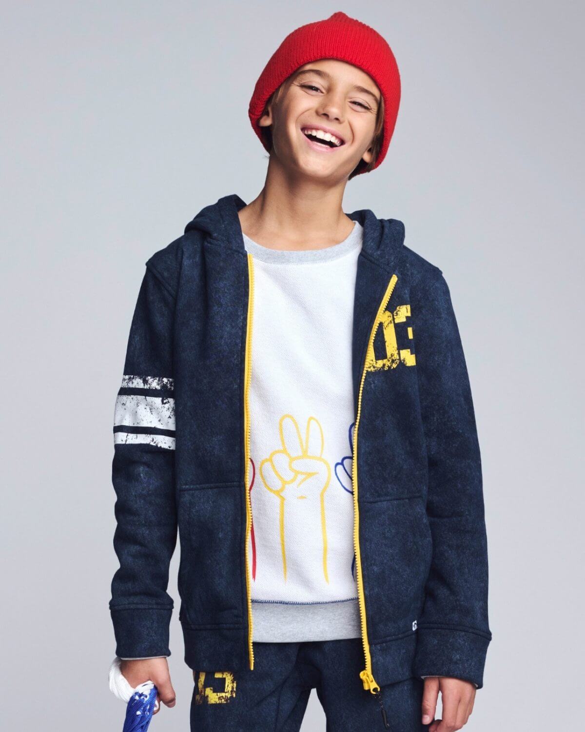 Child wearing a navy blue hoodie with yellow accents, white t-shirt with graphics, and red beanie on a gray background