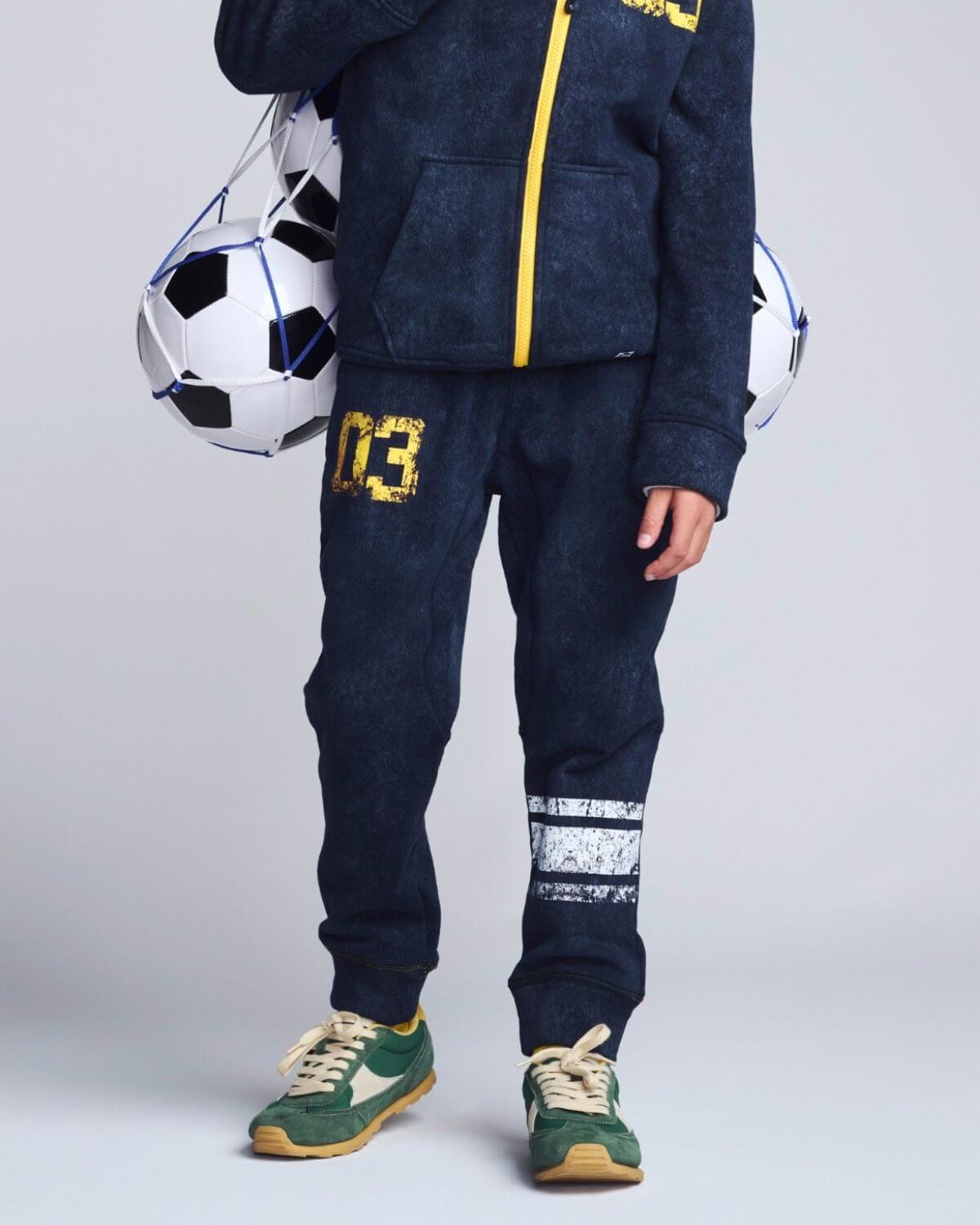 Person wearing navy tracksuit with yellow accents, holding a soccer ball on a light gray background
