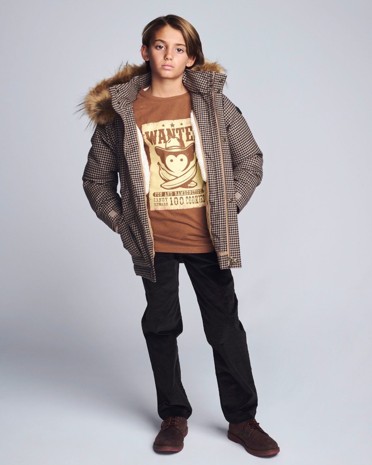 Person wearing a checkered coat with fur hood, brown t-shirt, and black pants on a light gray background
