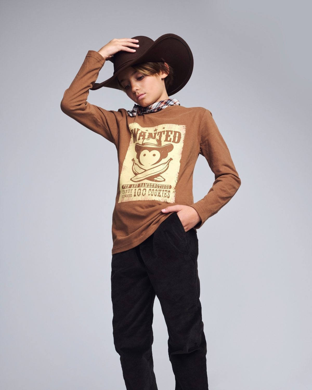 Person wearing a brown long-sleeve shirt with a graphic design, black pants, and a wide-brimmed hat on a gray background.