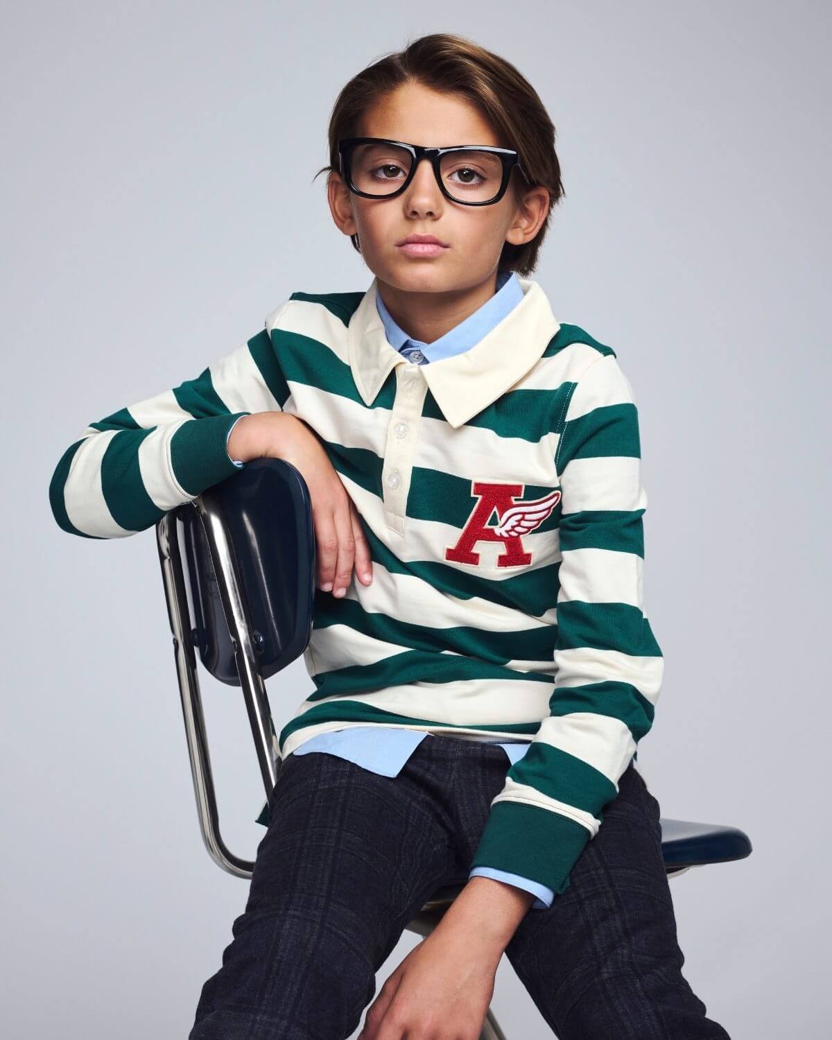 Child wearing a green and white striped sweater with a logo, sitting on a chair against a plain background