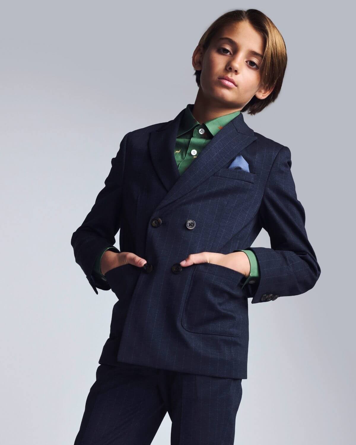 Young boy wearing a navy suit with a green shirt and blue pocket square on a light gray background