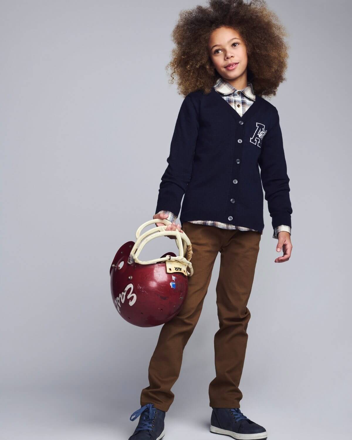 Child wearing a navy cardigan with a logo, holding a red helmet against a gray background