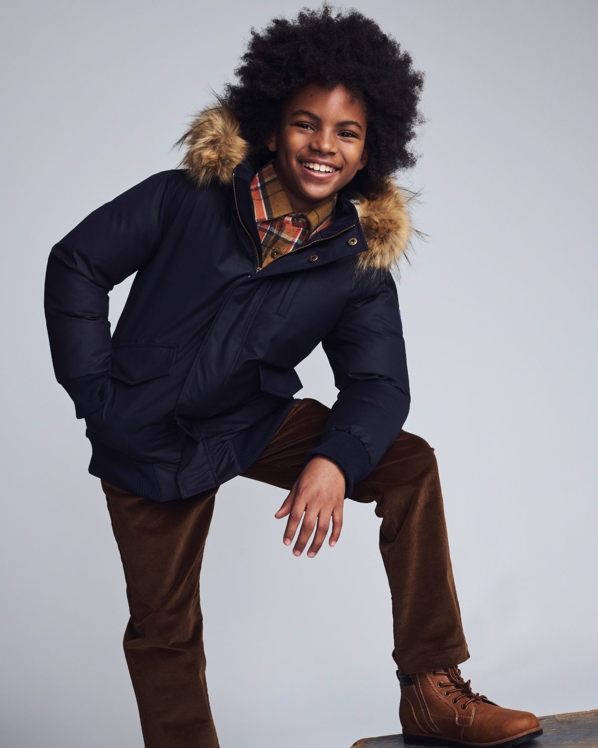 Child wearing a dark blue coat with fur hood, plaid shirt, and brown pants on a gray background