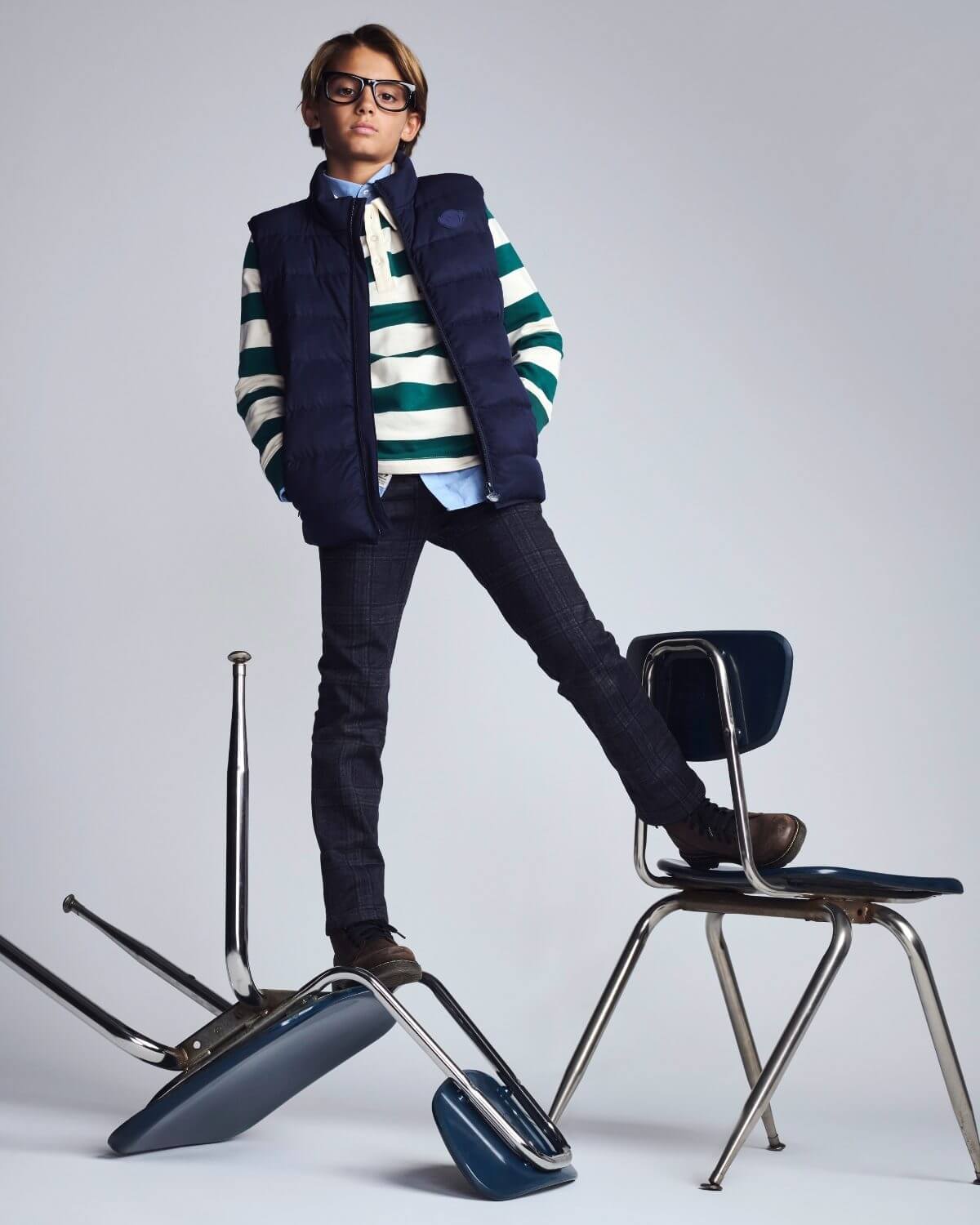 Person wearing a striped sweater and vest with a backpack, standing on a chair against a plain background