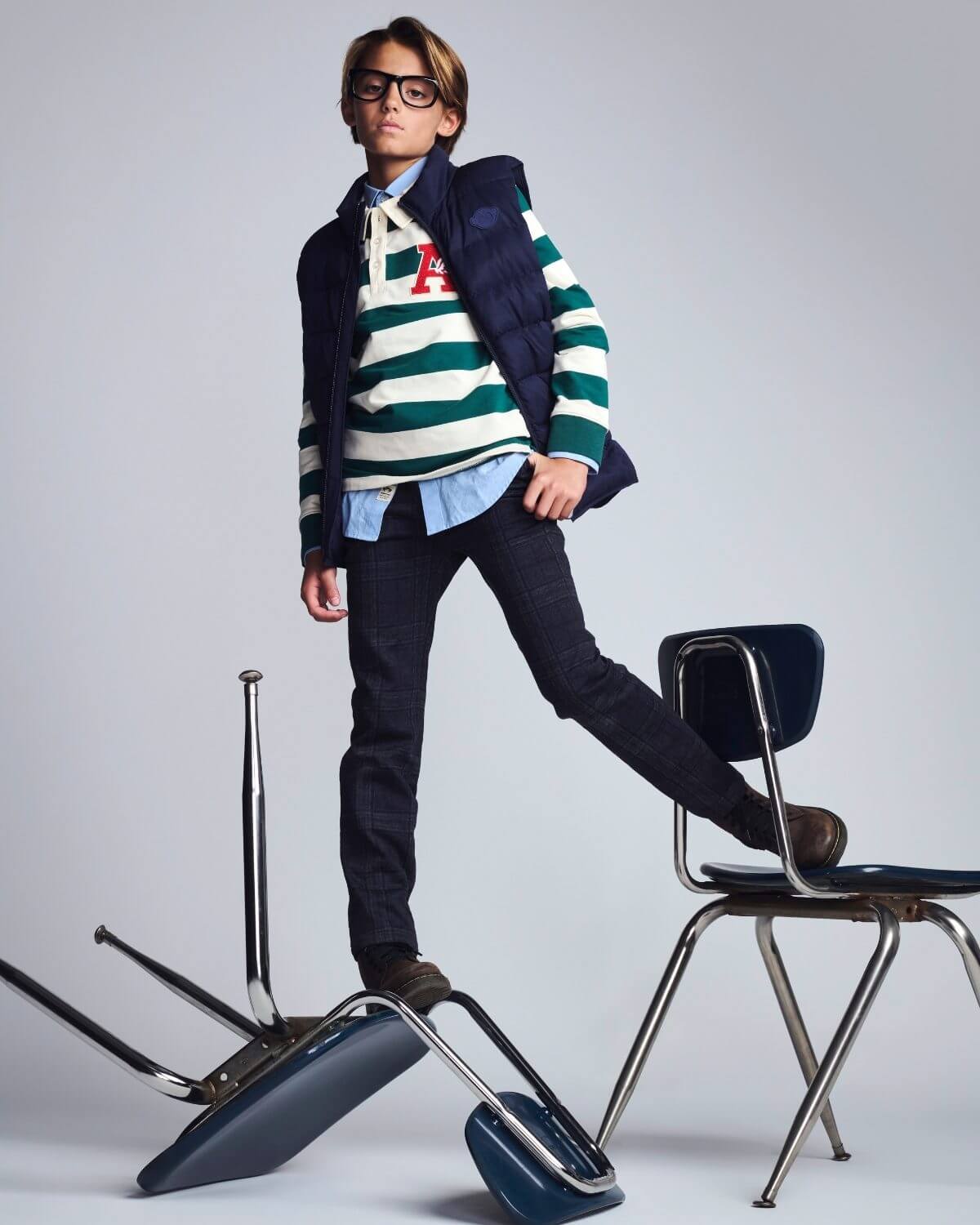 Person wearing a striped sweater and vest with a backpack, standing on a chair against a plain background