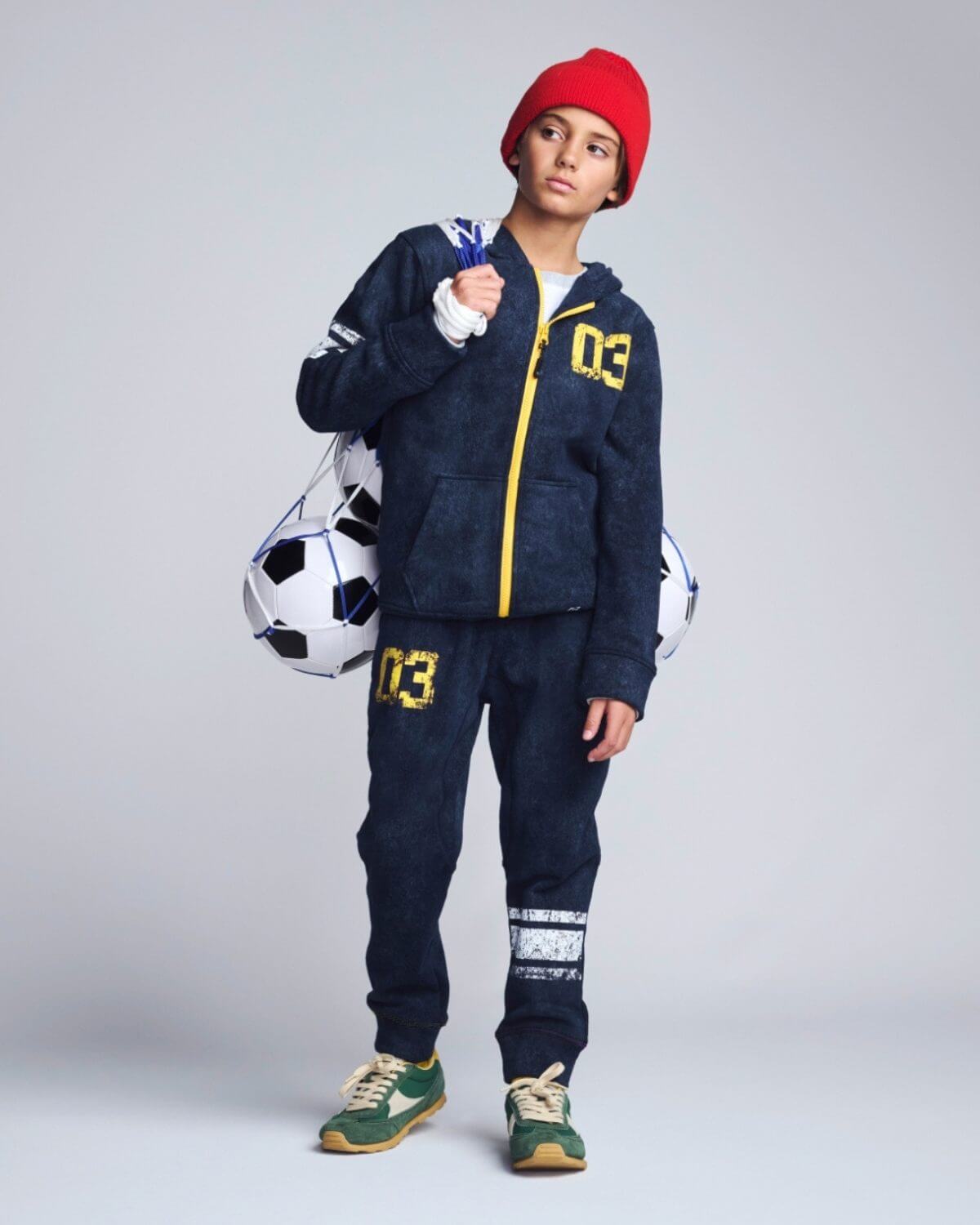 Person wearing a navy tracksuit with yellow accents, holding a soccer ball, on a light gray background