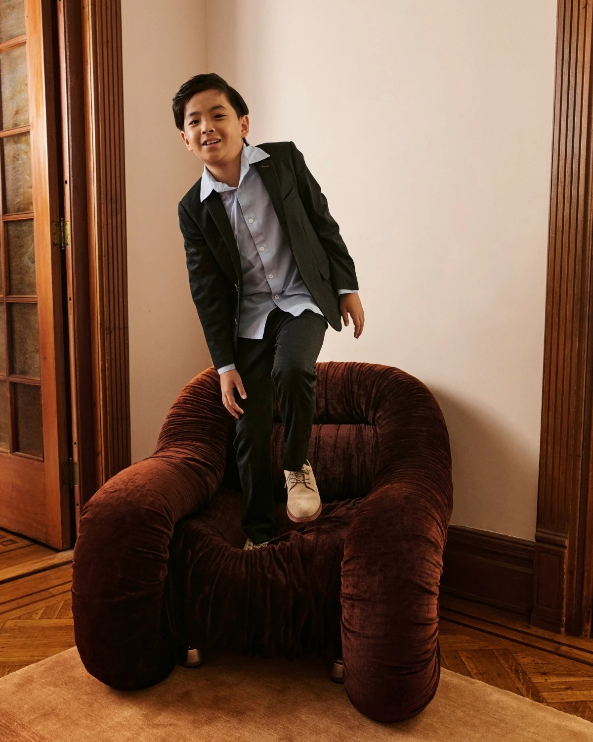 A boy wearing a navy suit, seated in a velvet chair, alternate view. Appaman Fall/Winter 2026 collection.